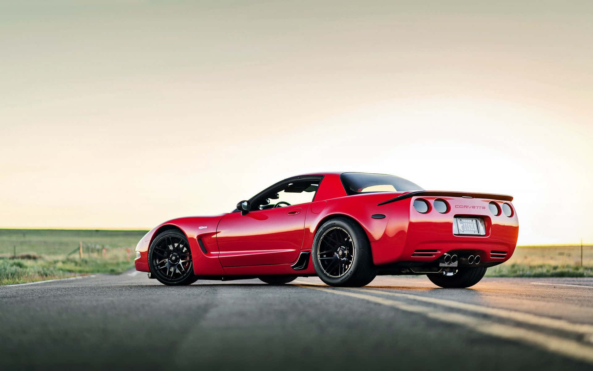 corvette 桌布 壁紙 — 一輛鮮紅色的Corvette跑車在黃金時段沿著鄉村柏油路滑行，柔和陽光突顯其流線曲線和黑色車輪。