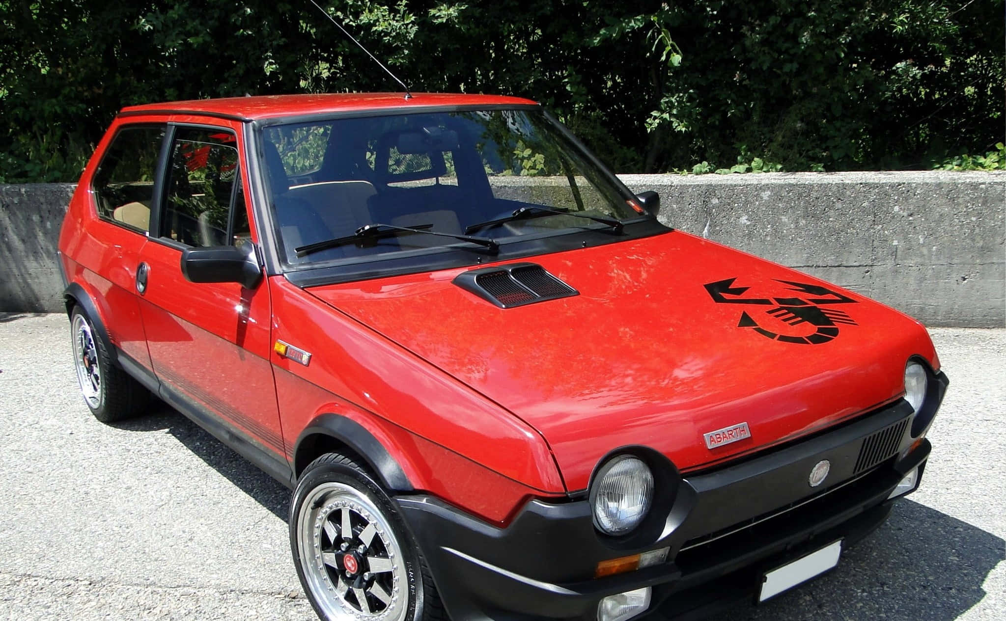 Fiat Ritmo 桌布 壁紙 — 一輛鮮紅色的Fiat 126p，帶有黑色飾邊和蠍子貼紙，停在陽光普照下的碎石路上，旁邊是石牆。