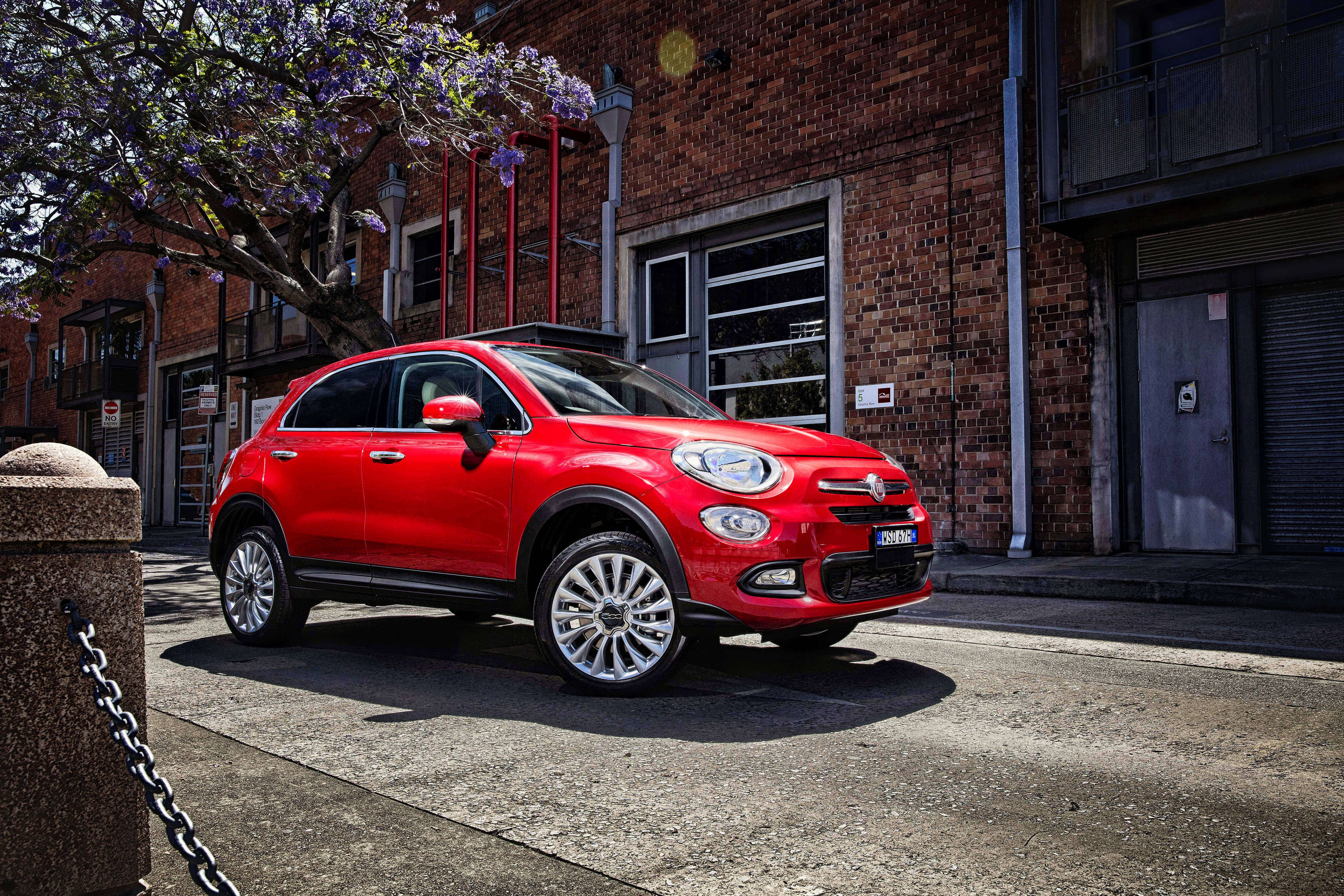 Fiat 桌布 壁紙 — 一輛鮮紅色的Fiat 500X SUV停放在陽光明媚的城市街道上，旁邊有開花的樹和磚造建築。