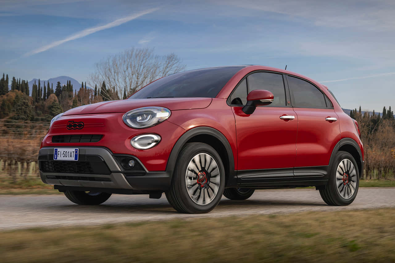 Fiat 桌布 壁紙 — 一輛鮮紅色的Fiat 500X SUV在鄉村道路上行駛，背景有模糊的綠化和遠處的山脈，天空清澈湛藍。