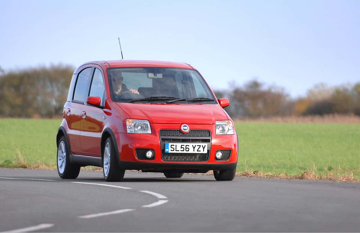 Fiat Panda 桌布 壁紙 — 一輛鮮紅色的Fiat Panda在晴朗的藍天下沿著蜿蜒的鄉村道路行駛，兩旁是綠色的田野。