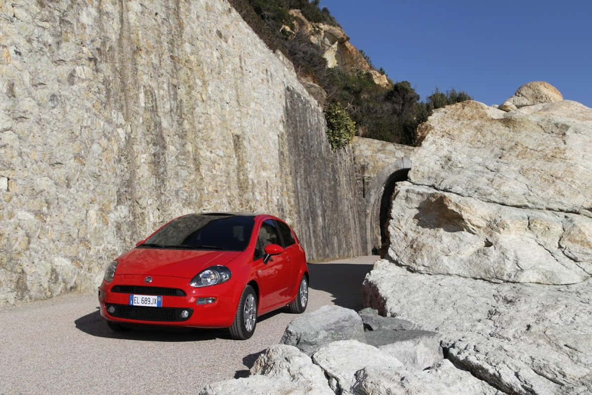 Fiat Punto 桌布 壁紙 — 一輛鮮紅色的Fiat Punto在晴朗藍天下，沿著陽光明媚的碎石路行駛，兩旁是高聳崎嶇的石崖。