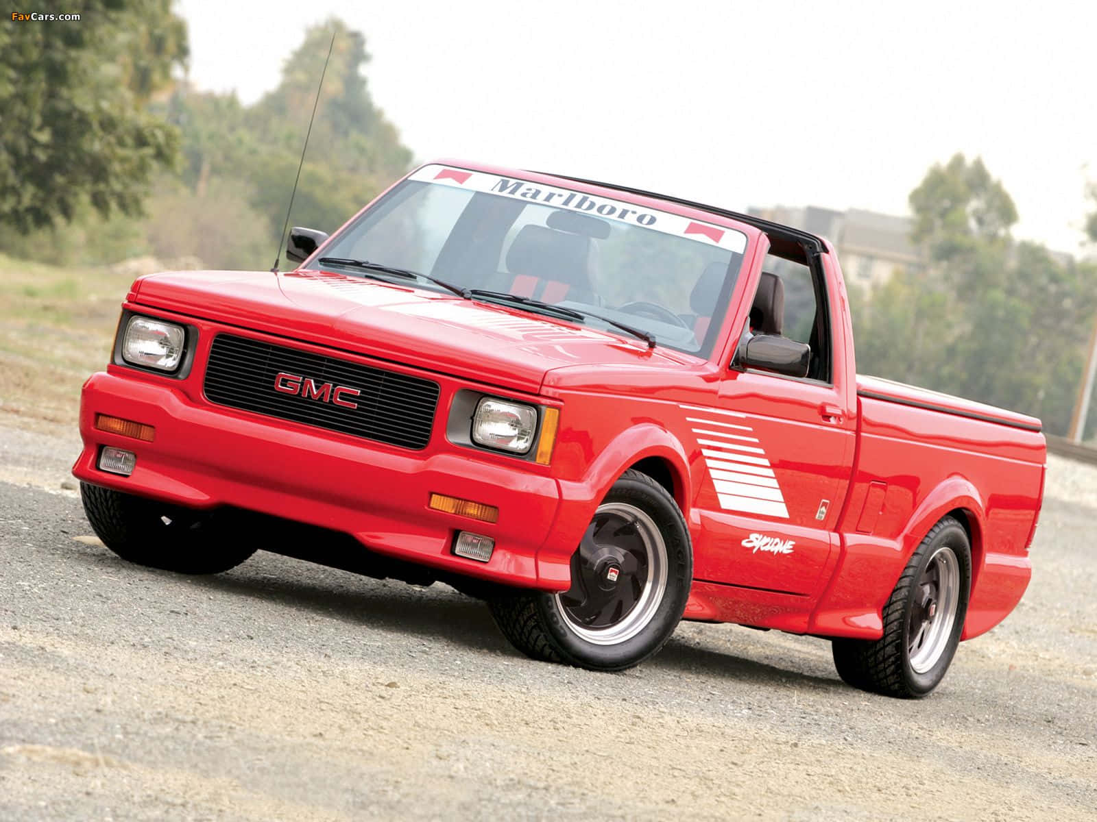 Gmc Syclone 桌布 壁紙 — 一輛鮮紅色的GMC Syclone皮卡車，帶有賽車貼紙，停在樹木環繞的碎石路上。
