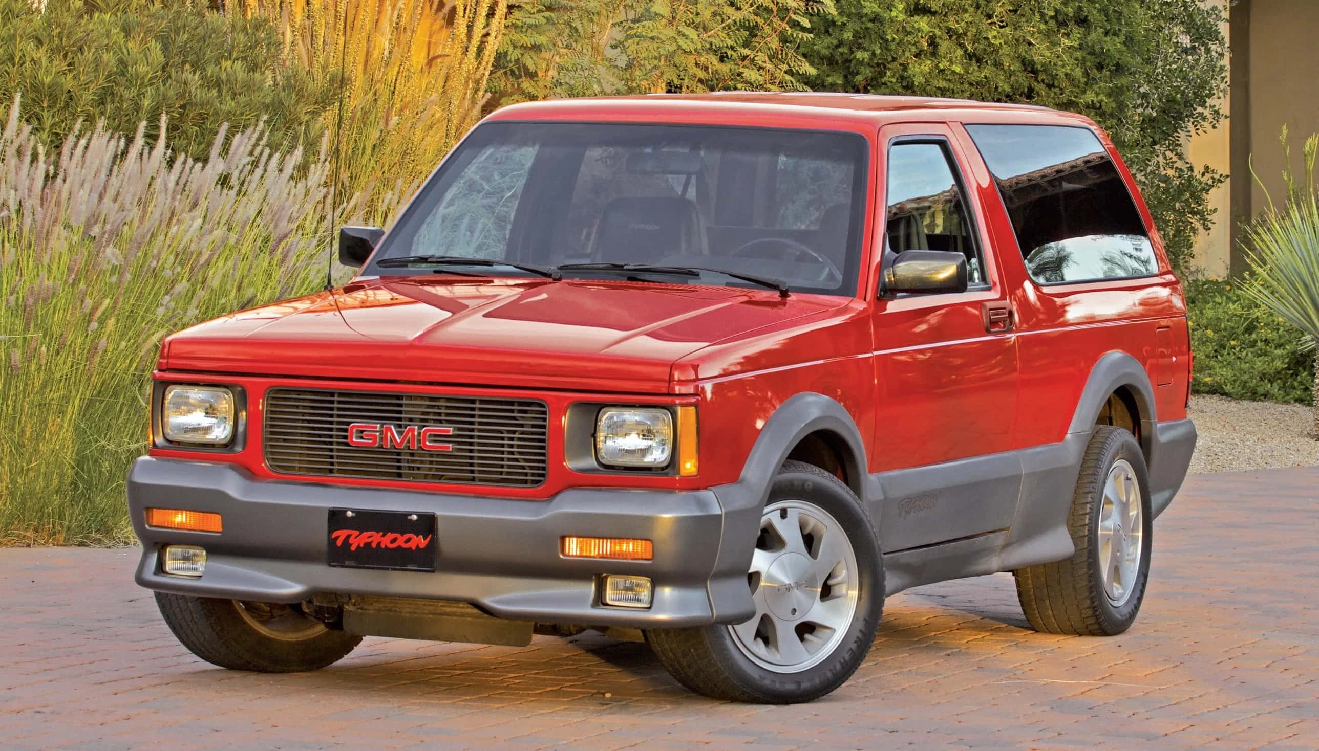 Gmc Typhoon 桌布 壁紙 — 一輛鮮紅色的GMC Typhoon SUV停放在磚砌車道上，背景有高草和樹木。