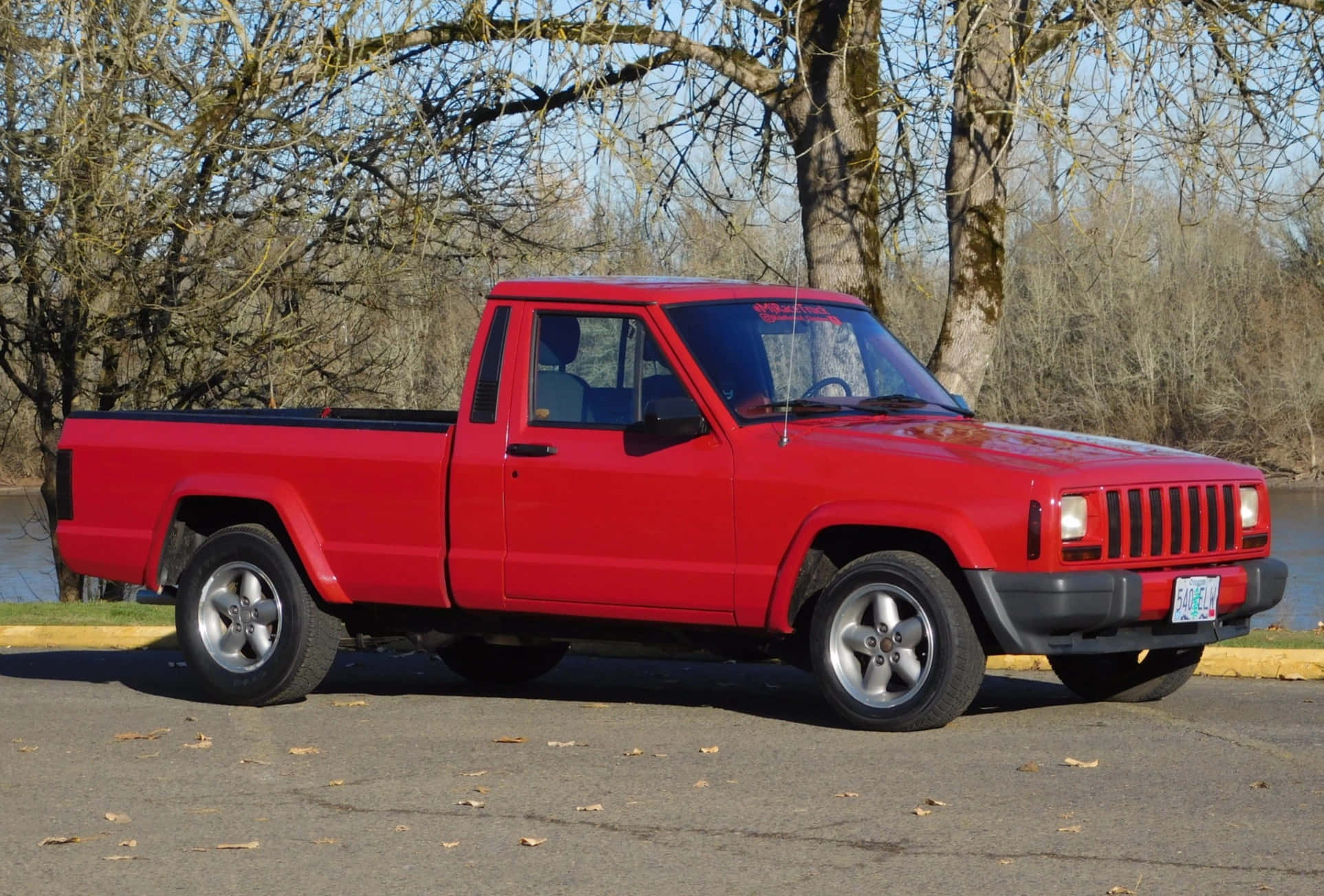 Jeep Comanche 桌布 壁紙 — 一輛鮮紅色的Jeep Cherokee皮卡車停在瀝青路上，旁邊是一條河，河邊有光禿禿的樹木，天空清澈湛藍。
