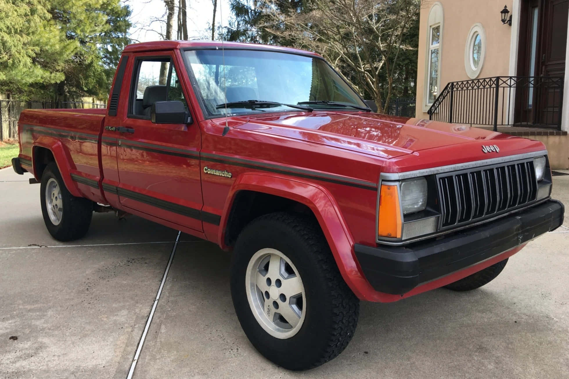 Jeep Comanche 桌布 壁紙 — 一輛鮮紅色的Jeep Comanche皮卡車停在房子前面的混凝土車道上，周圍是光禿禿的樹木。