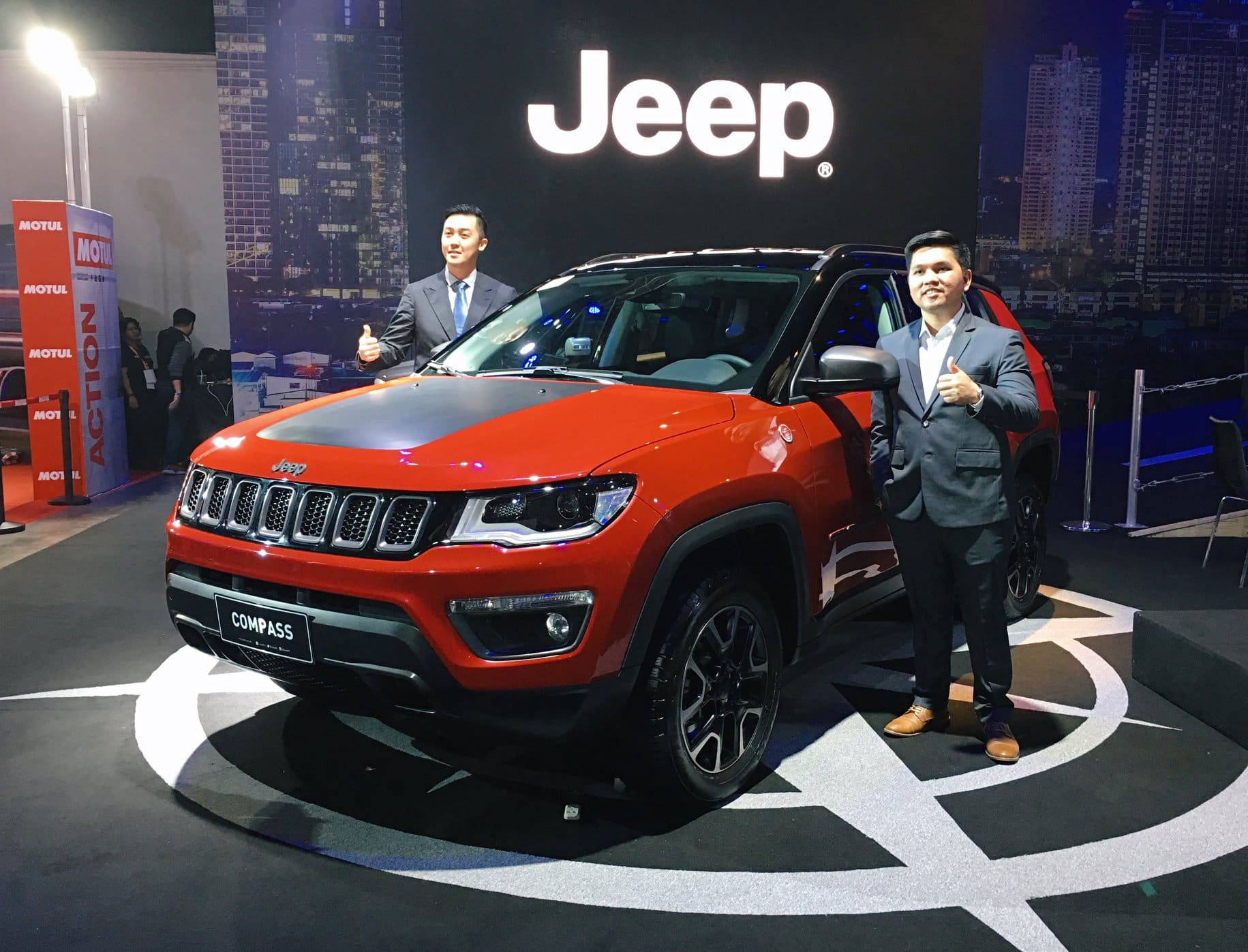 紅色美學動圖 桌布 壁紙 — 一輛鮮紅色的Jeep Compass SUV在車展的圓形平台上展示，旁邊有兩名穿西裝的男子豎起大拇指。