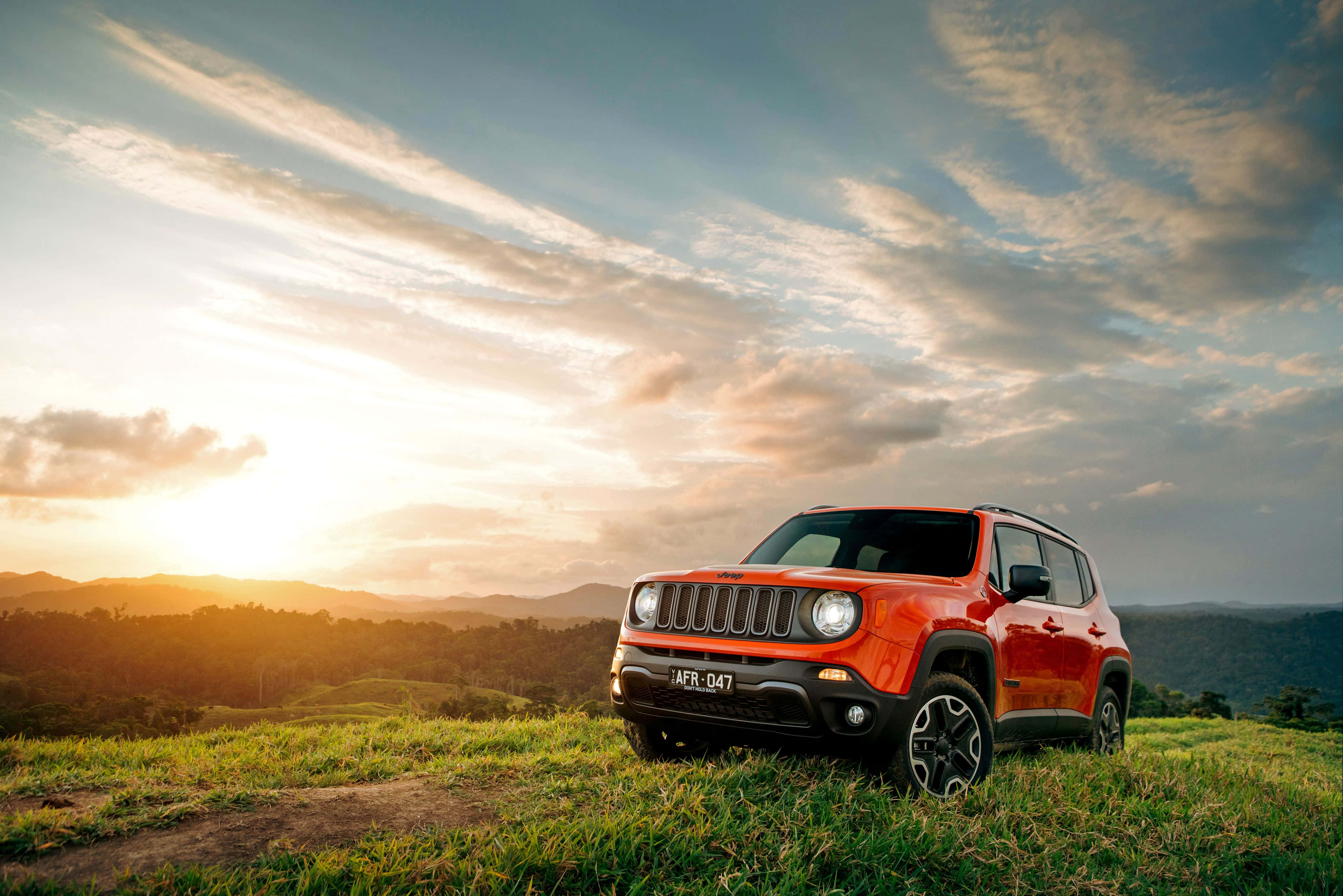Jeep Renegade 桌布 壁紙 — 一輛鮮紅色的Jeep Renegade在日落時分停泊在長滿草的山丘上，金色的陽光映照著起伏的丘陵和散落的雲朵。