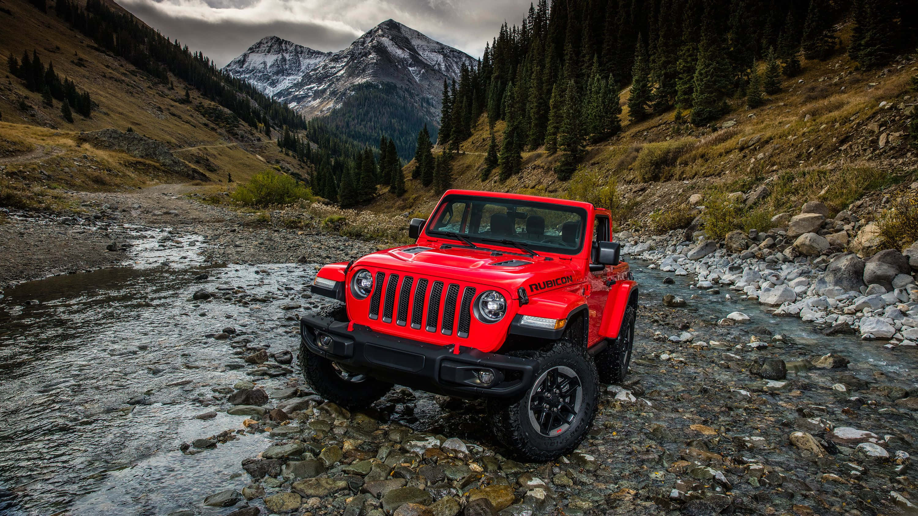 吉普 桌布 壁紙 — 一輛鮮紅色的Jeep Wrangler Rubicon在陰天背景下，穿越岩石密佈的山間溪流，背景是白雪覆蓋的山峰。