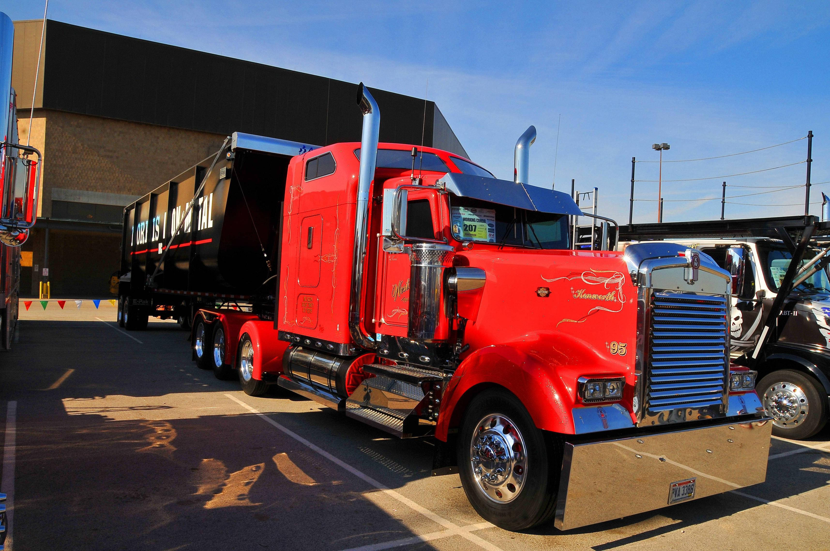 Kenworth 桌布 壁紙 — 一輛鮮紅色的Kenworth半掛卡車，帶有鍍鉻飾邊和定制塗裝，停在晴朗藍天下的停車場。