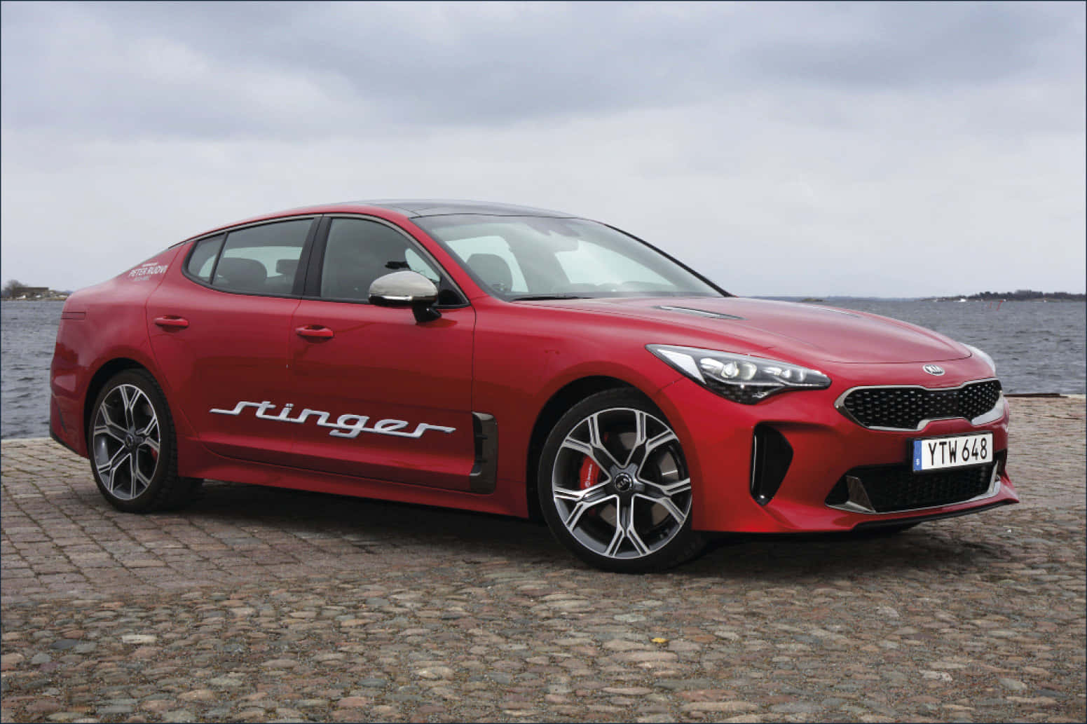 Kia Stinger 桌布 壁紙 — 一輛鮮紅色的Kia Stinger運動轎車停泊在鵝卵石碼頭上，俯瞰著陰天下一片平靜的灰色海洋。
