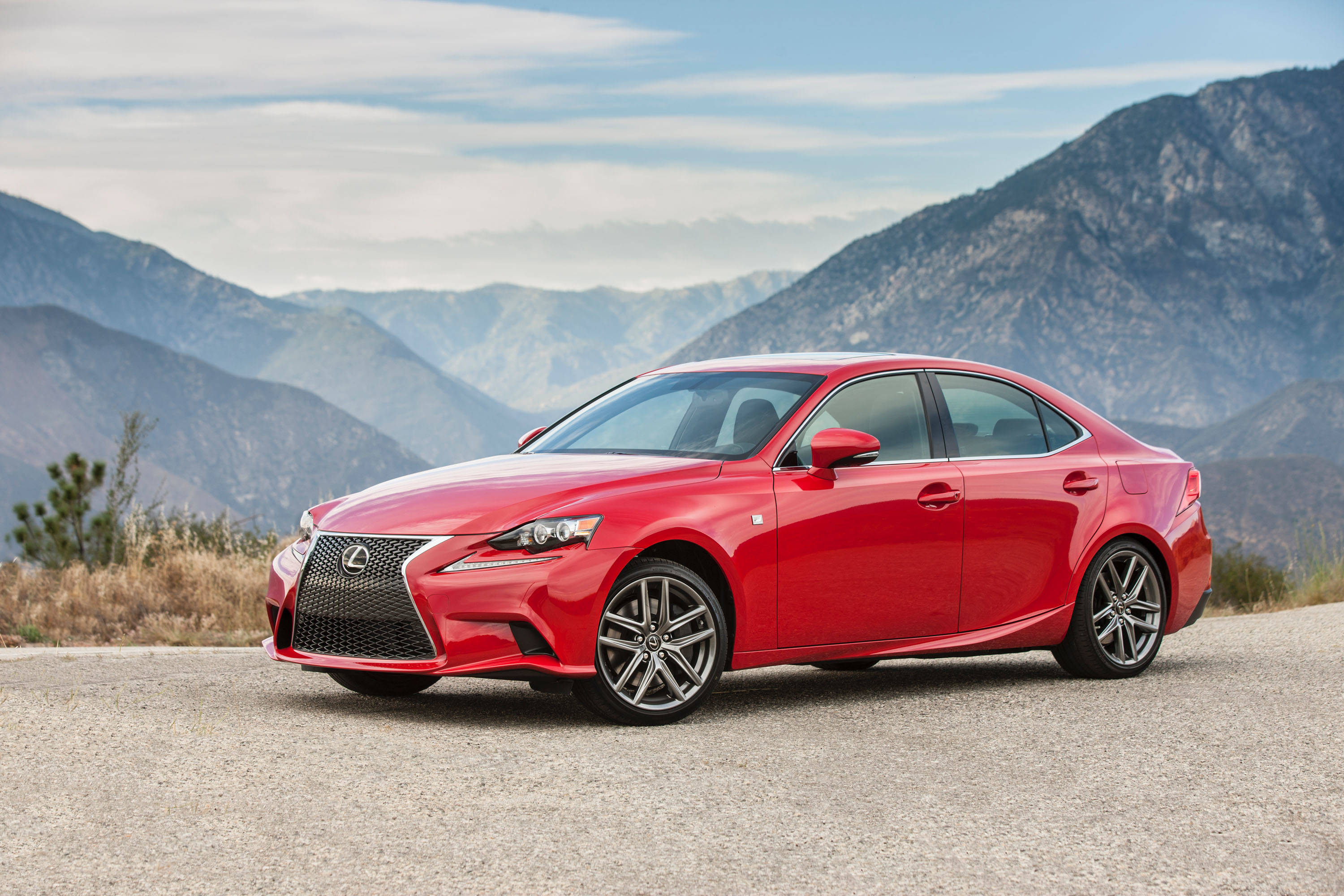 lexus iPhone 桌布 壁紙 — 一輛鮮紅色的Lexus房車在碎石路上閃閃發光，背後是崎嶇的山脈，在柔和的藍天下聳立。