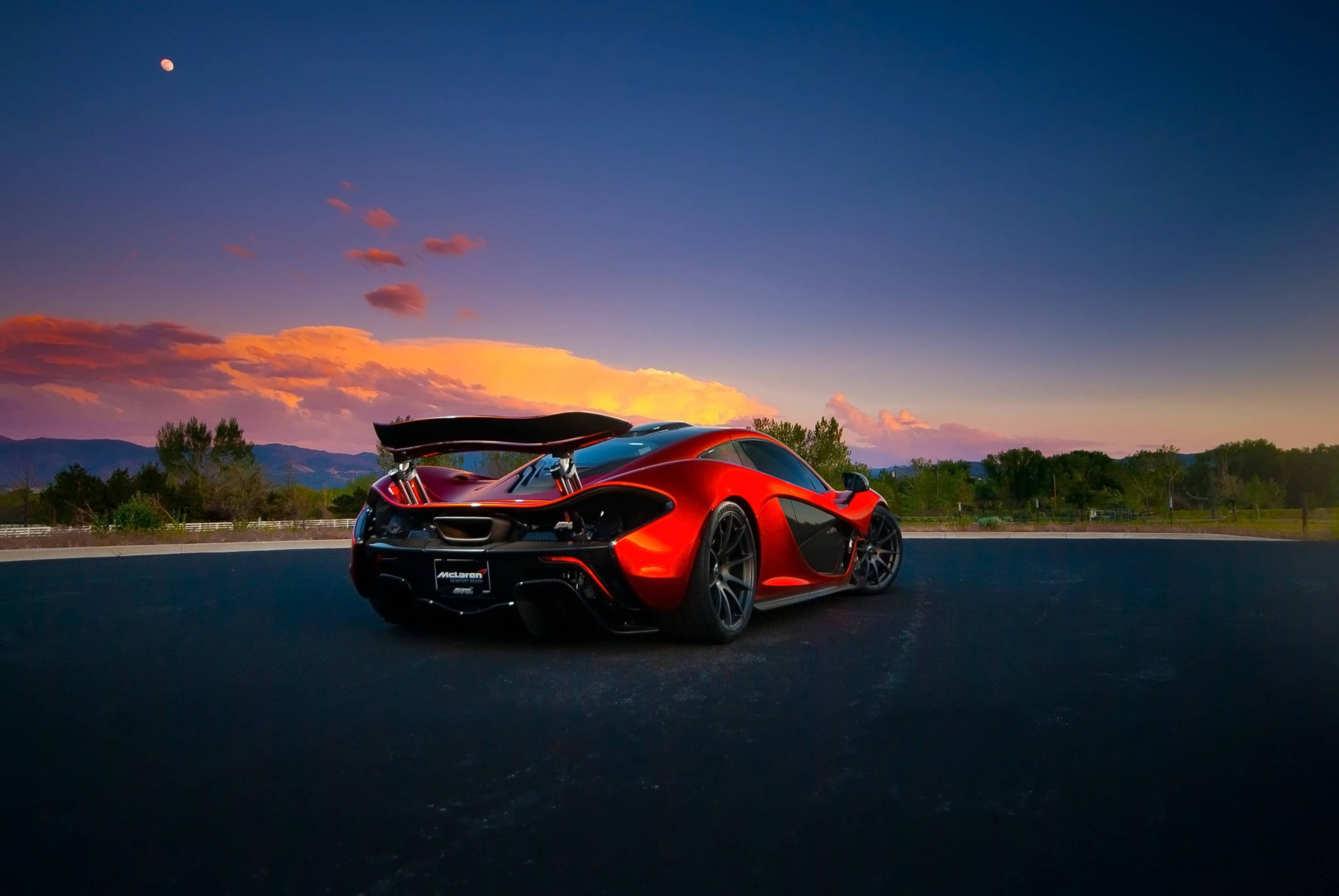 跑車 桌布 壁紙 — 一輛鮮紅色的McLaren跑車，尾翼升起，停在日落時分的深色瀝青路上，背景是遠山和色彩繽紛的天空，還有一輪可見的月亮。