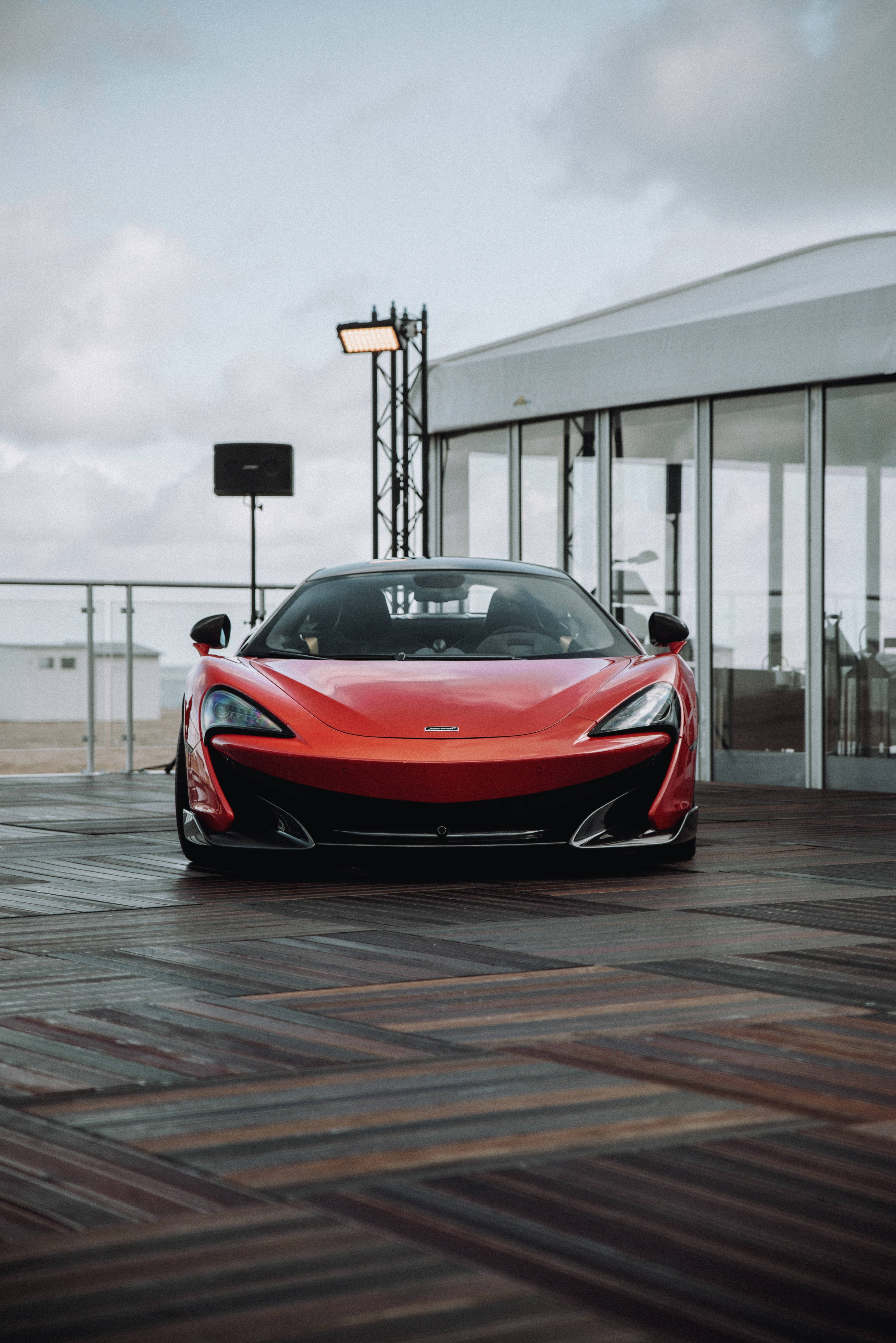 麥拉倫 桌布 壁紙 — 一輛鮮紅色的McLaren跑車停在現代玻璃建築附近的木質甲板上，天空多雲。