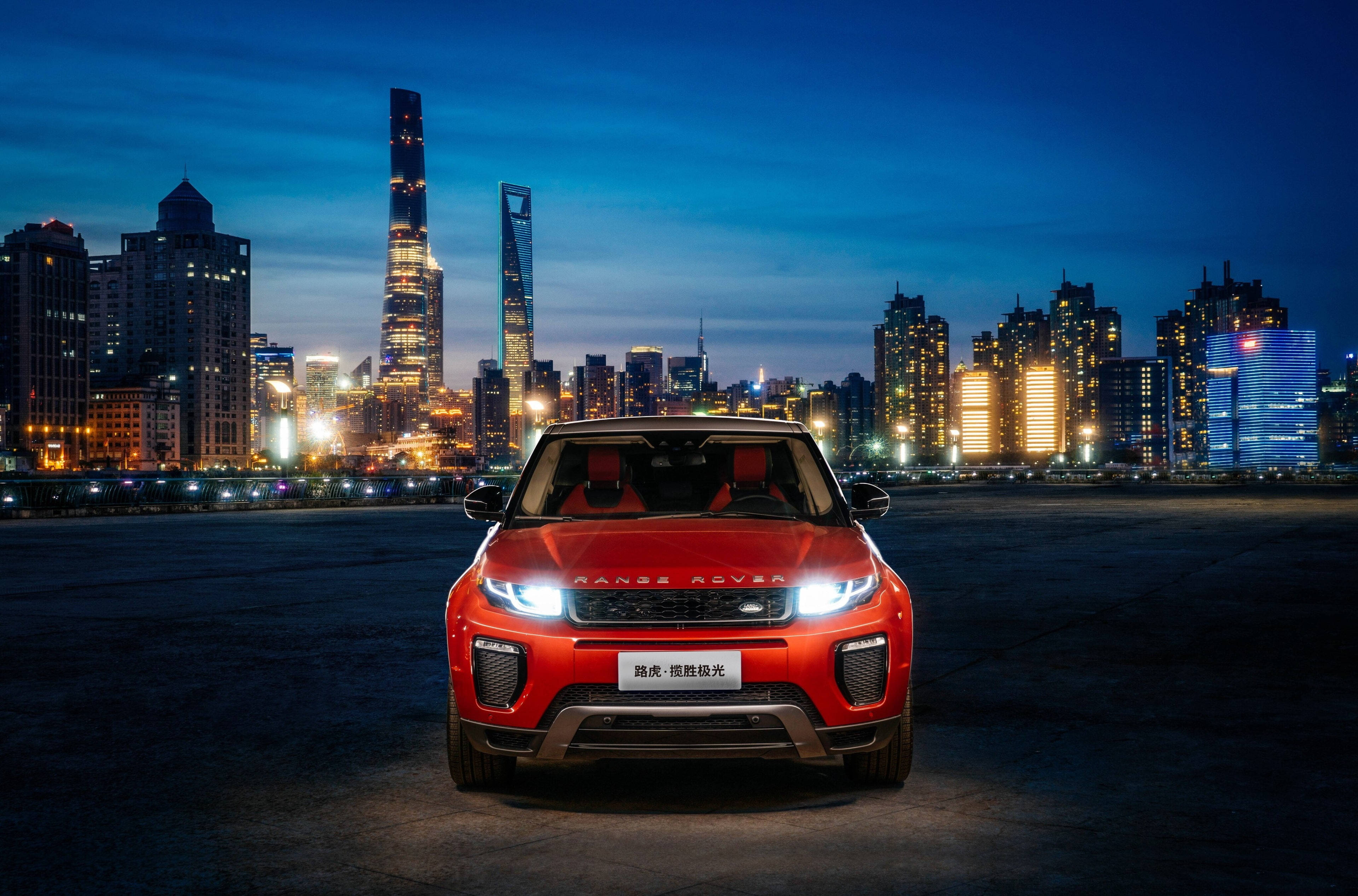 高清 桌布 壁紙 — 一輛鮮紅色的Range Rover Evoque在暮色中，以燈火通明的摩天大樓為背景，在城市燈光下閃閃發光。