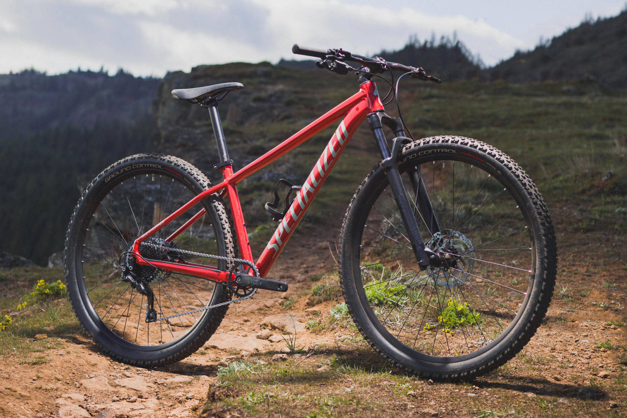 腳踏車 桌布 壁紙 — 一輛鮮紅色的Specialized登山車在崎嶇的泥土小徑上蓄勢待發，背景是連綿起伏的綠色丘陵和遠處的森林覆蓋山脈，天空多雲。
