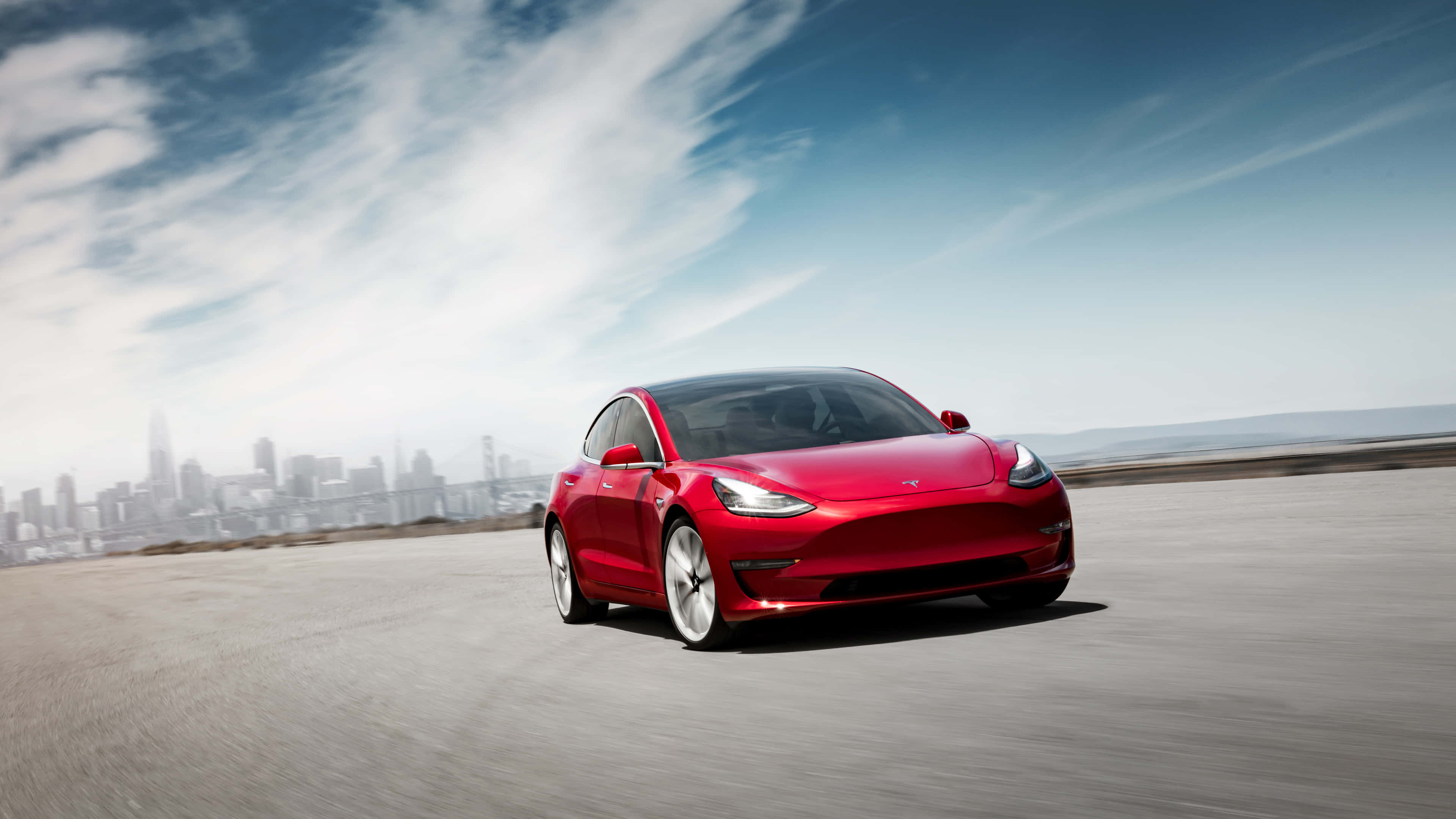 Tesla Model 3 桌布 壁紙 — 一輛鮮紅色的Tesla Model 3在部分多雲的藍天下，沿著一條鋪砌的道路疾馳，背景是朦朧的城市天際線。