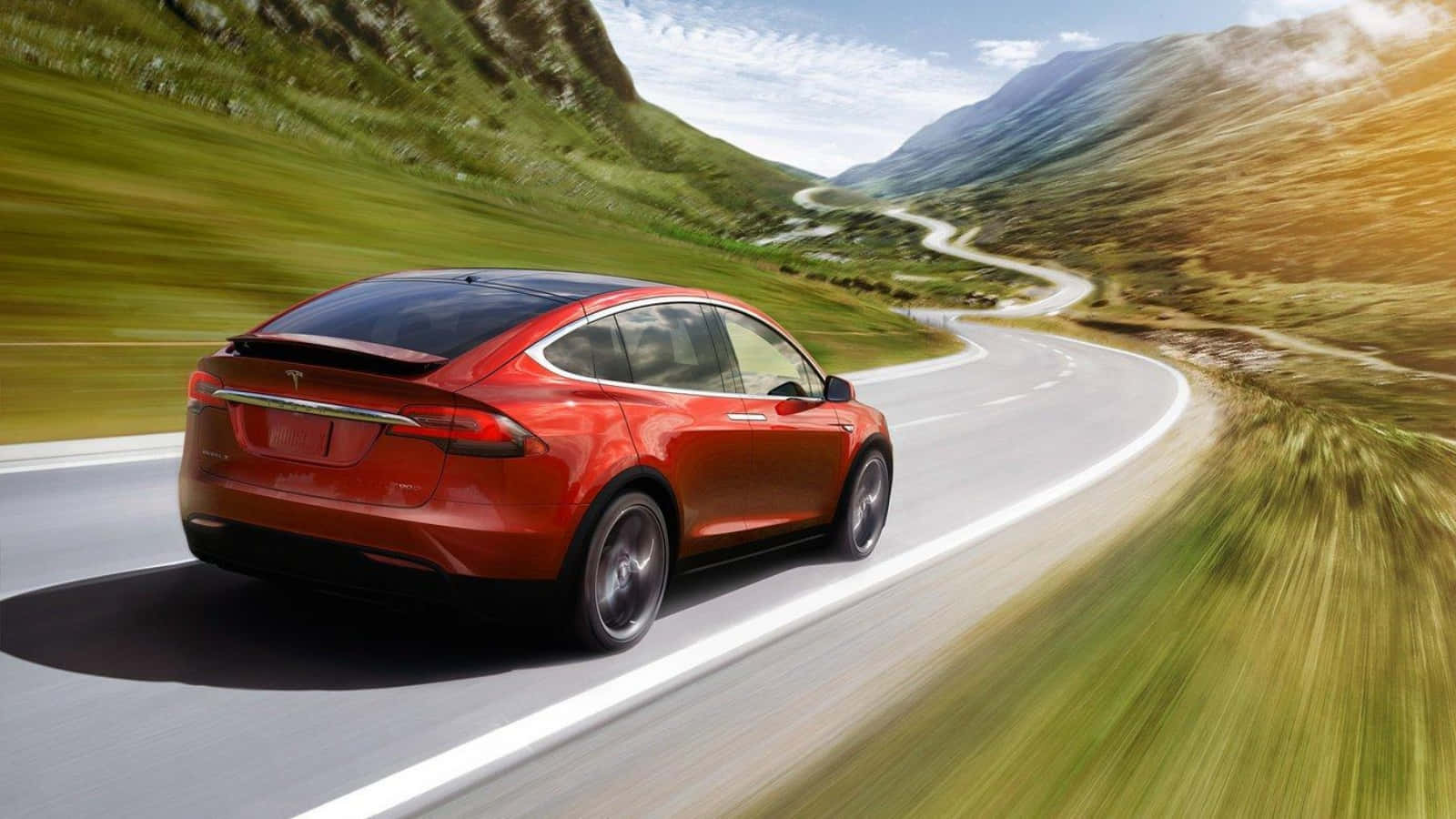 Tesla Model 3 桌布 壁紙 — 一輛鮮紅色的Tesla Model X在晴朗藍天下，沿著蜿蜒的山路穿過翠綠的山丘飛馳。