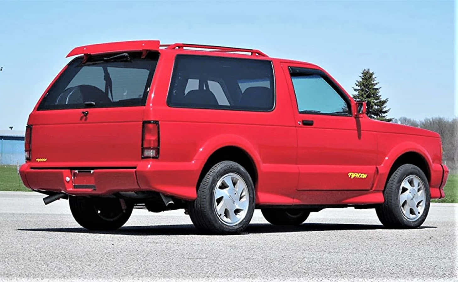 Gmc Typhoon 桌布 壁紙 — 一輛鮮紅色的Typhoon SUV停在瀝青路面上，背景是清澈的藍天和樹木。