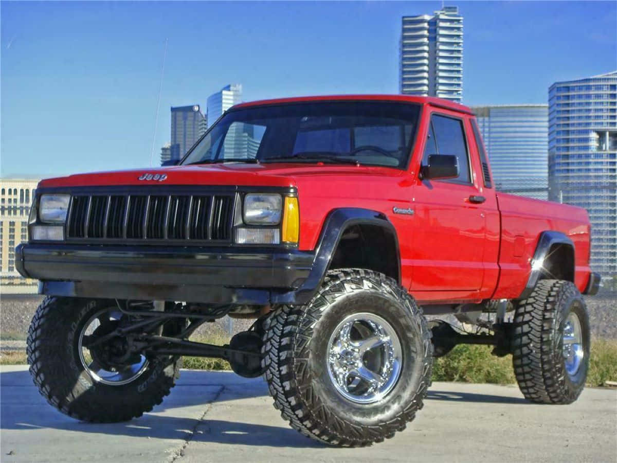Jeep Comanche 桌布 壁紙 — 一輛鮮紅色的升高Jeep Cherokee皮卡車，配備超大輪胎，在晴朗藍天下傲然矗立於現代城市摩天大樓的背景前。