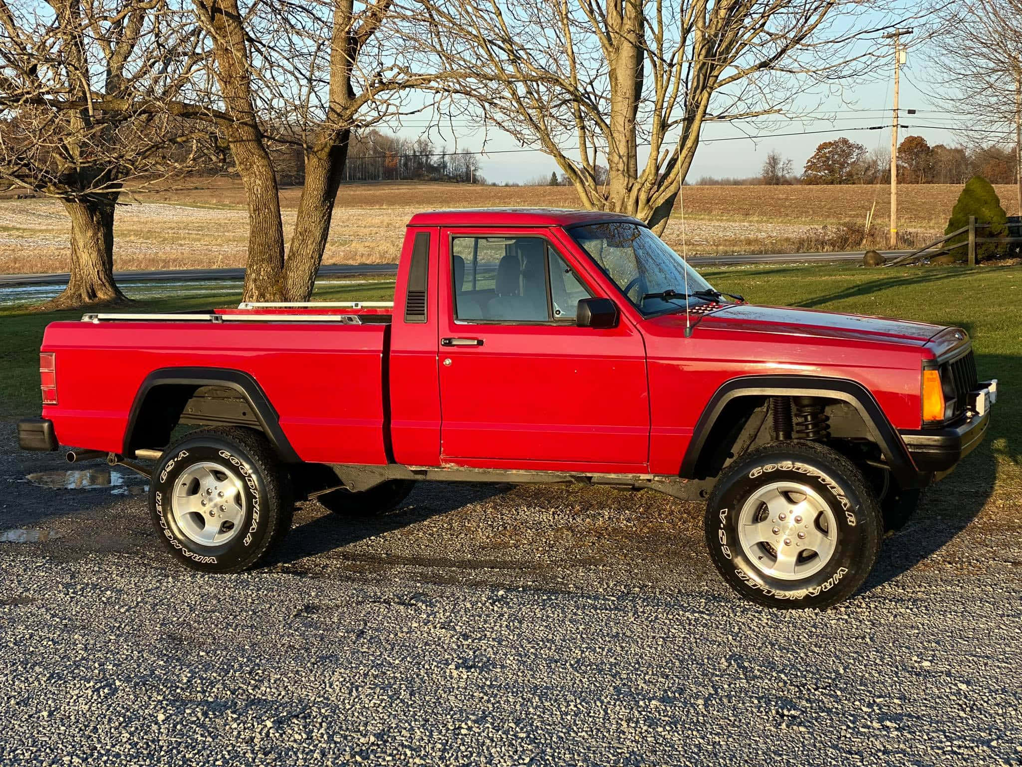 Jeep Comanche 桌布 壁紙 — 一輛鮮紅色的升高皮卡車停在碎石上，背景是光禿禿的樹木和開闊的田野，天空晴朗。
