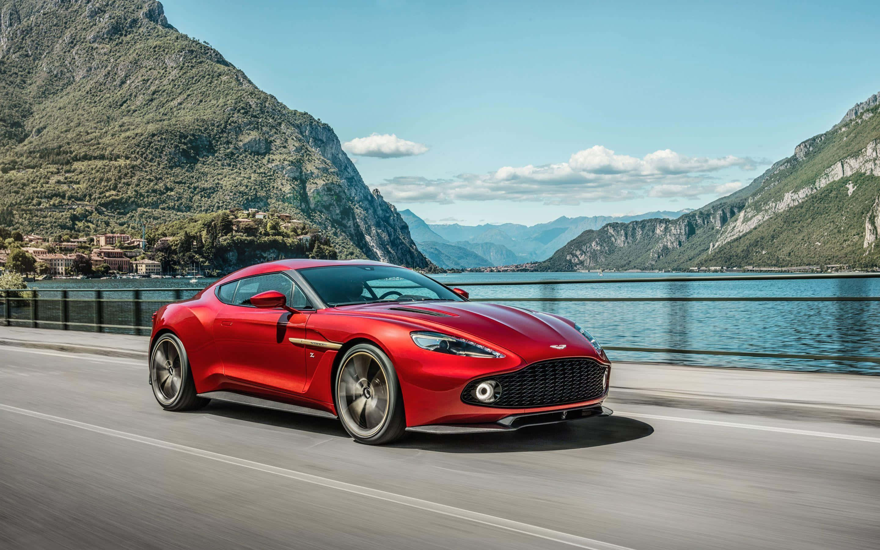 Aston Martin Vanquish 桌布 壁紙 — 一輛鮮紅色的奧斯頓馬丁在晴朗藍天下，沿著兩旁高聳翠綠山巒的湖邊道路飛馳。
