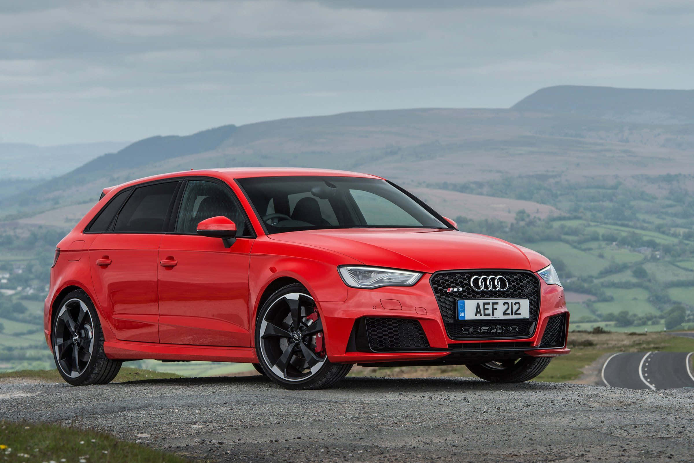 Audi Rs3 桌布 壁紙 — 一輛鮮紅色的奧迪RS3 Sportback停泊在碎石路上，背景是起伏的綠色山丘和陰天。