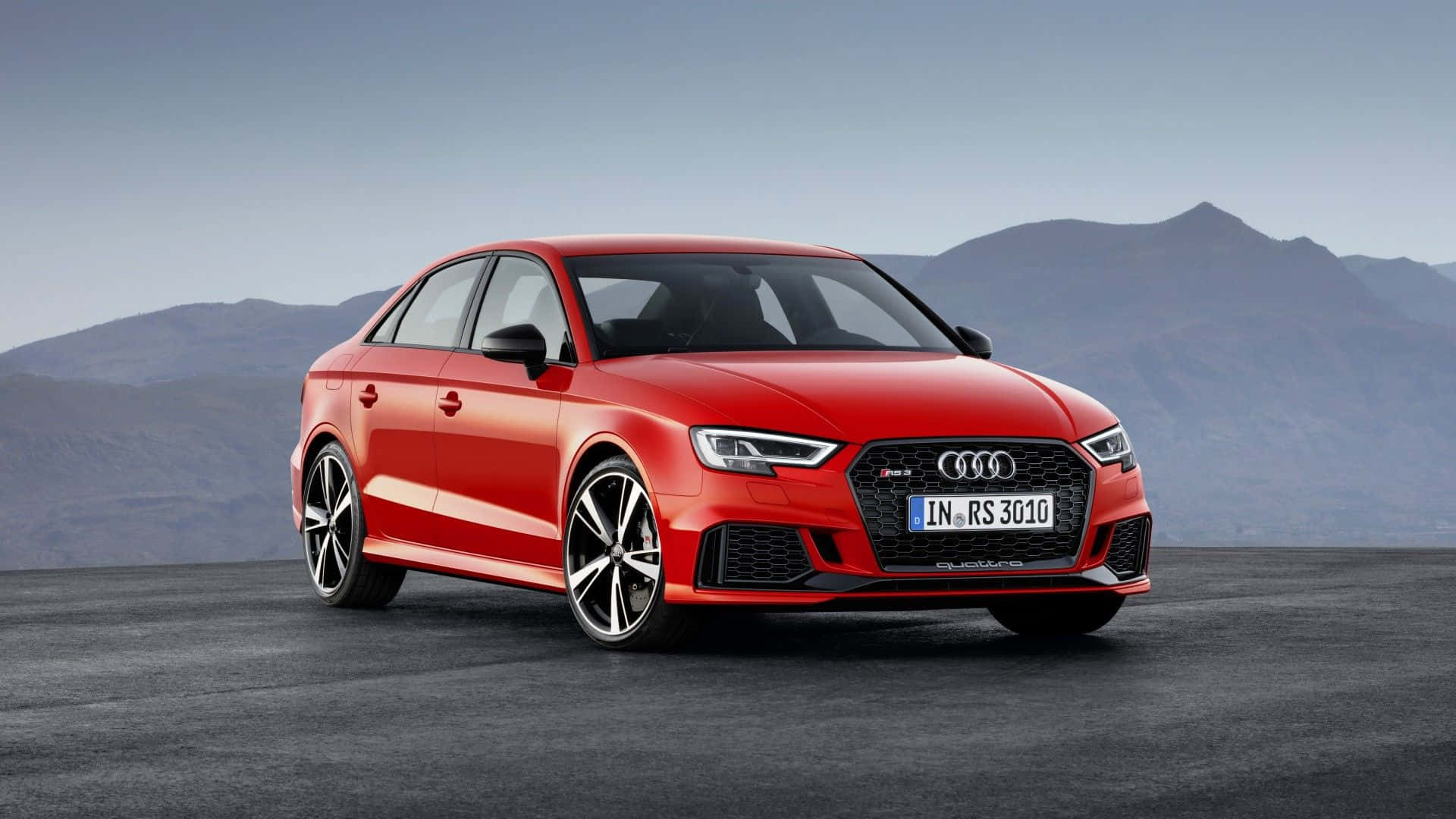 Audi Rs3 桌布 壁紙 — 一輛鮮紅色的奧迪RS3房車停在漆黑的瀝青路上，遠處有山脈，天空晴朗。