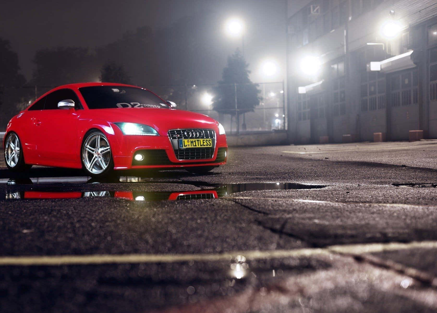 Audi Tt 桌布 壁紙 — 一輛鮮紅色的奧迪TT轎跑車在街燈下閃耀，其倒影在霧氣瀰漫的城市夜晚中，於雨水浸濕的水坑中閃爍。