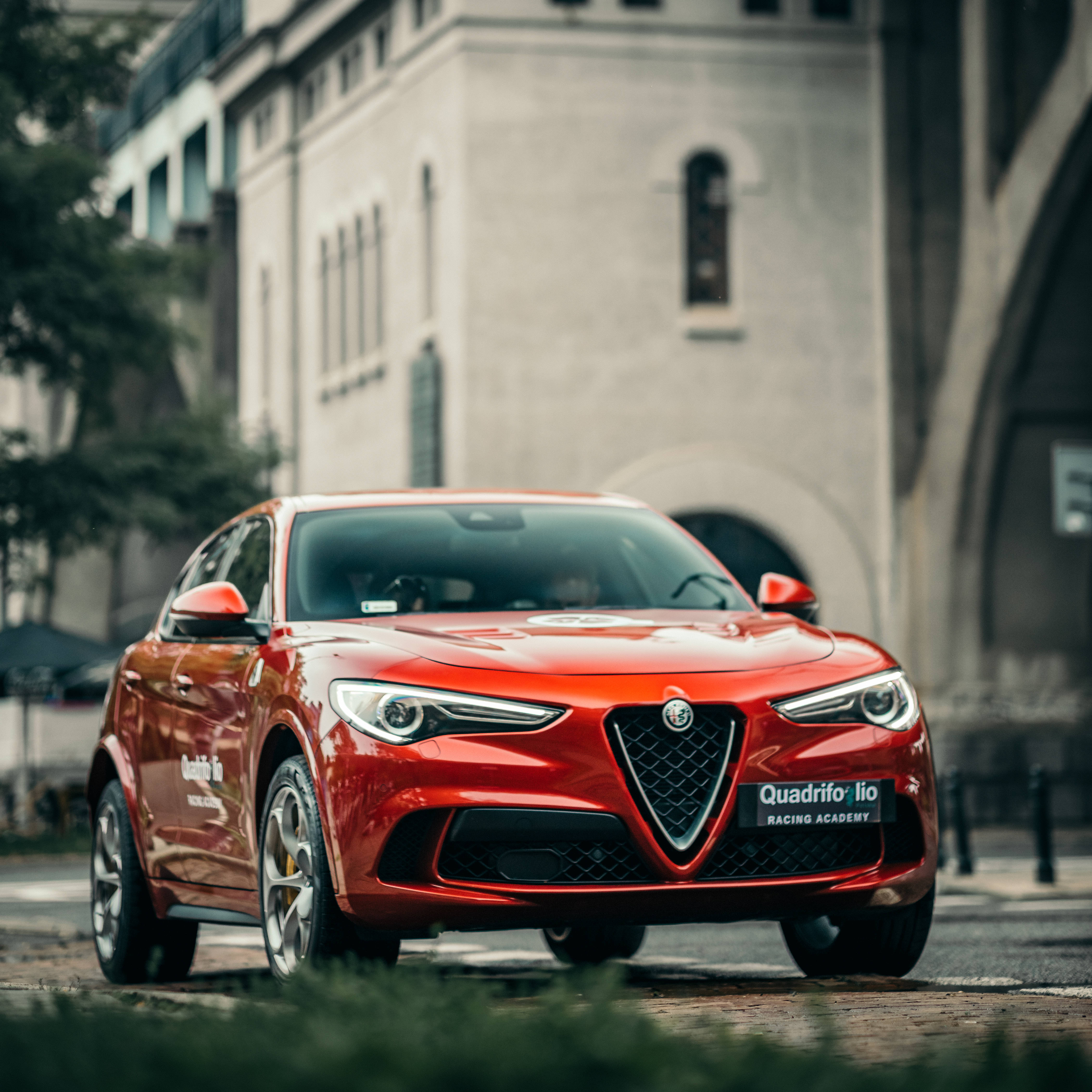 愛快羅密歐 桌布 壁紙 — 一輛鮮紅色的愛快羅密歐 Quadrifoglio 賽車停泊在歷史石建築前的鵝卵石街道上。
