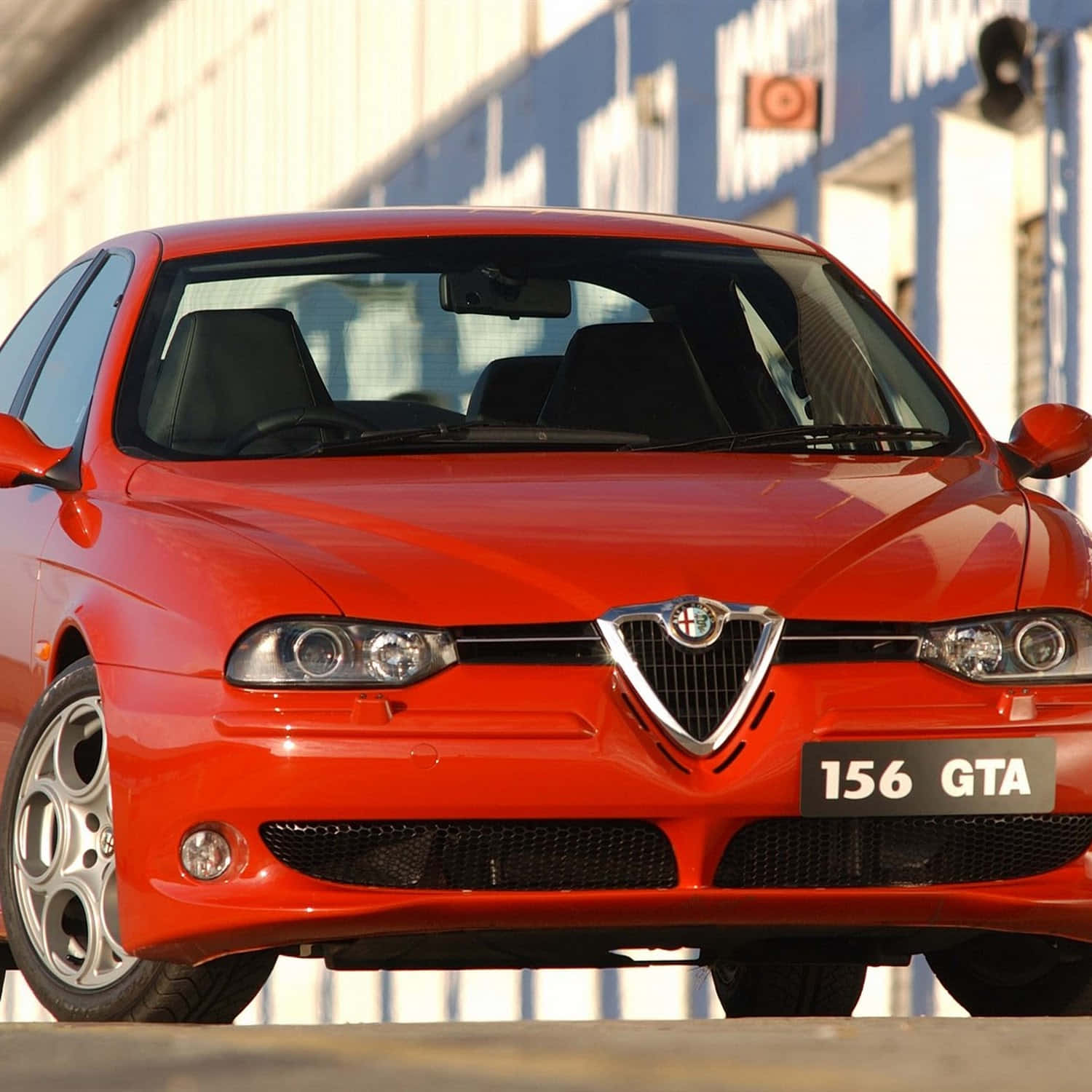 Alfa Romeo 156 桌布 壁紙 — 一輛鮮紅色的愛快羅密歐156 GTA跑車在明亮的陽光下閃閃發光，焦點集中在標誌性的水箱護罩和時尚的頭燈上。
