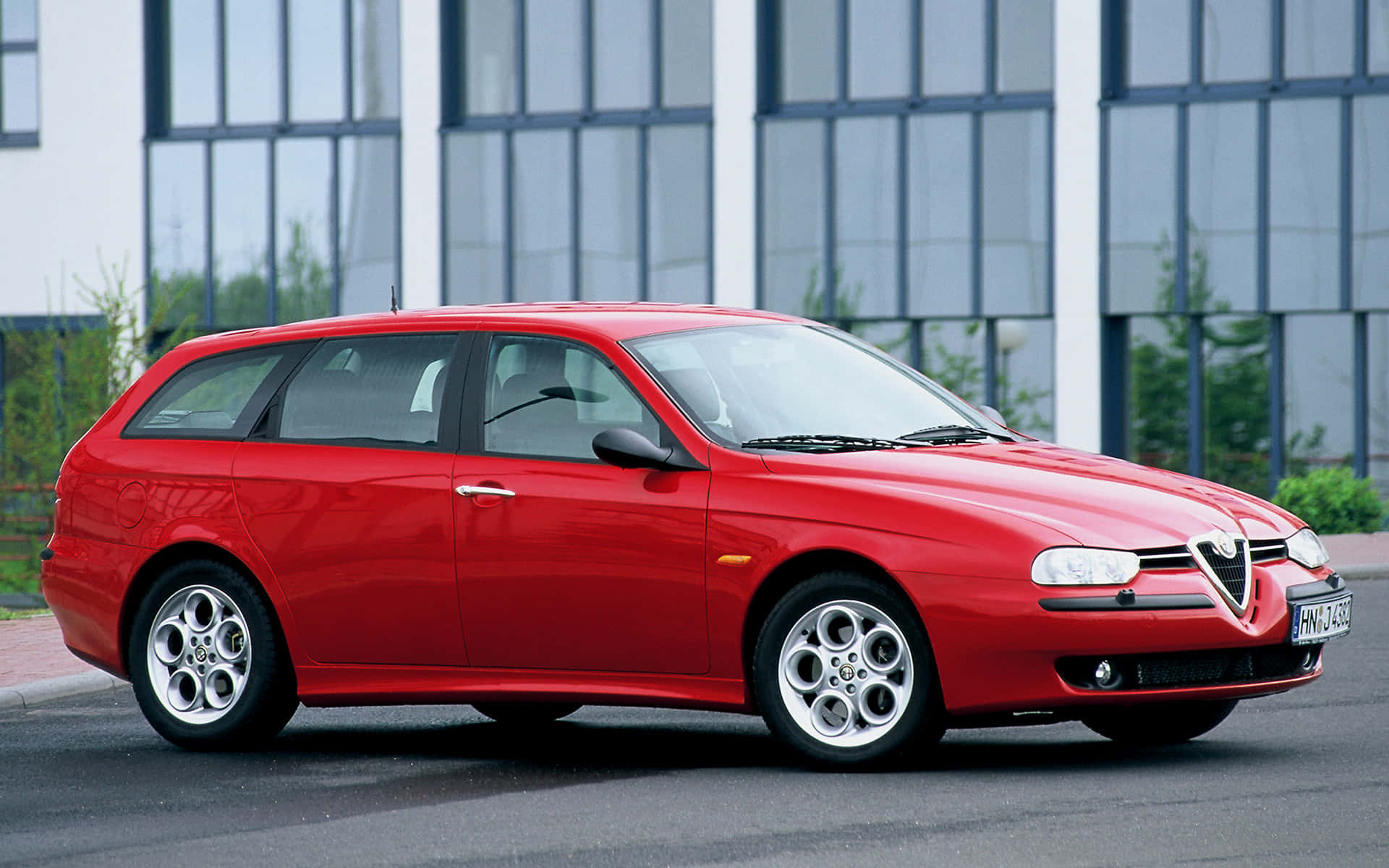 Alfa Romeo 156 桌布 壁紙 — 一輛鮮紅色的愛快羅密歐156旅行車停在現代玻璃建築前的瀝青路面上。