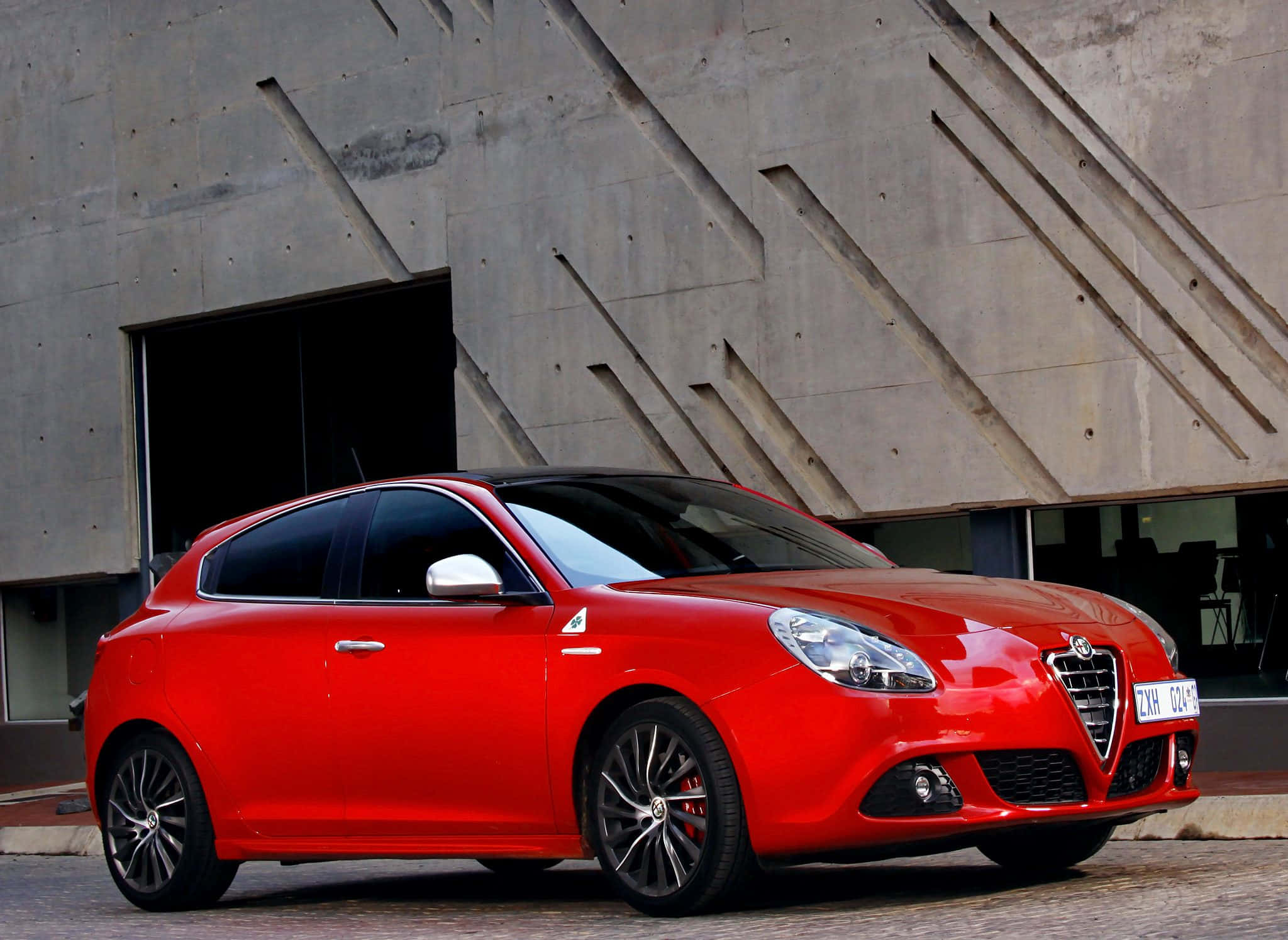 Alfa Romeo Giulietta 桌布 壁紙 — 一輛鮮紅色的愛快羅密歐Giulietta跑車停放在現代幾何線條的混凝土建築前。