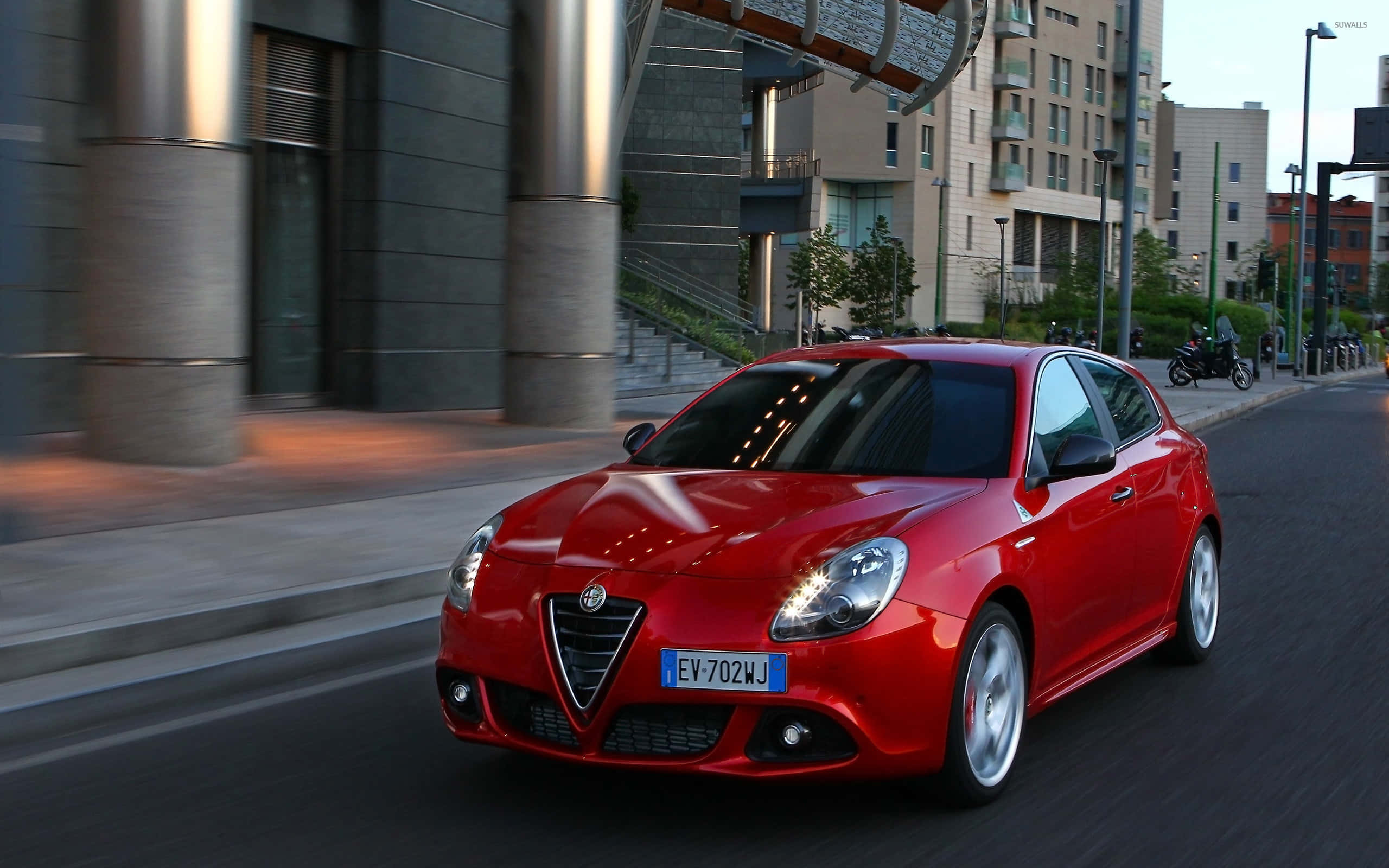 Alfa Romeo Giulietta 桌布 壁紙 — 一輛鮮紅色的愛快羅密歐Giulietta在黃昏時分沿著現代都市街道疾馳，背景帶有動態模糊效果。