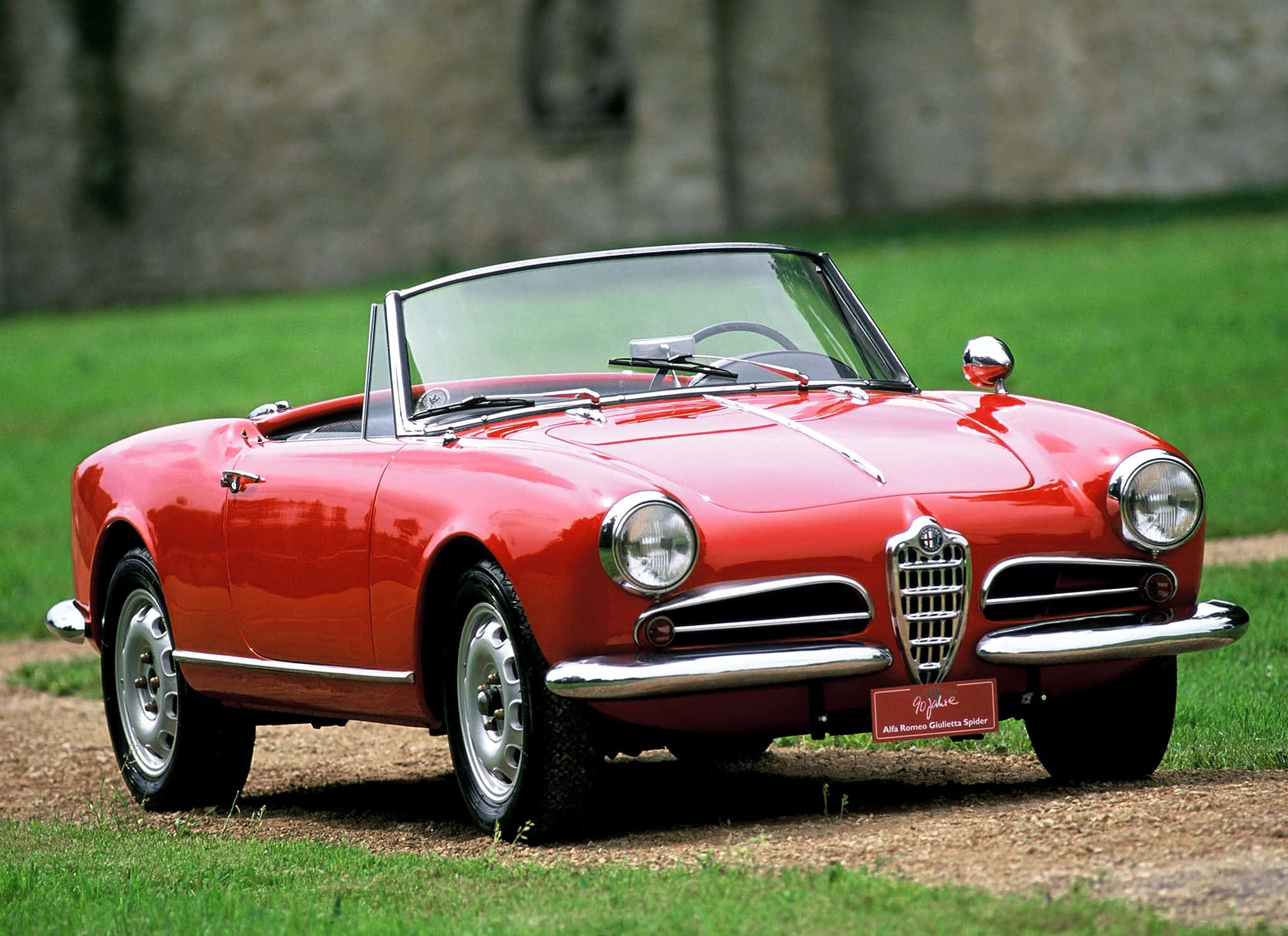 Alfa Romeo Spider 桌布 壁紙 — 一輛鮮紅色的愛快羅密歐Giulietta Spider敞篷車停在草地上，背景是石牆。