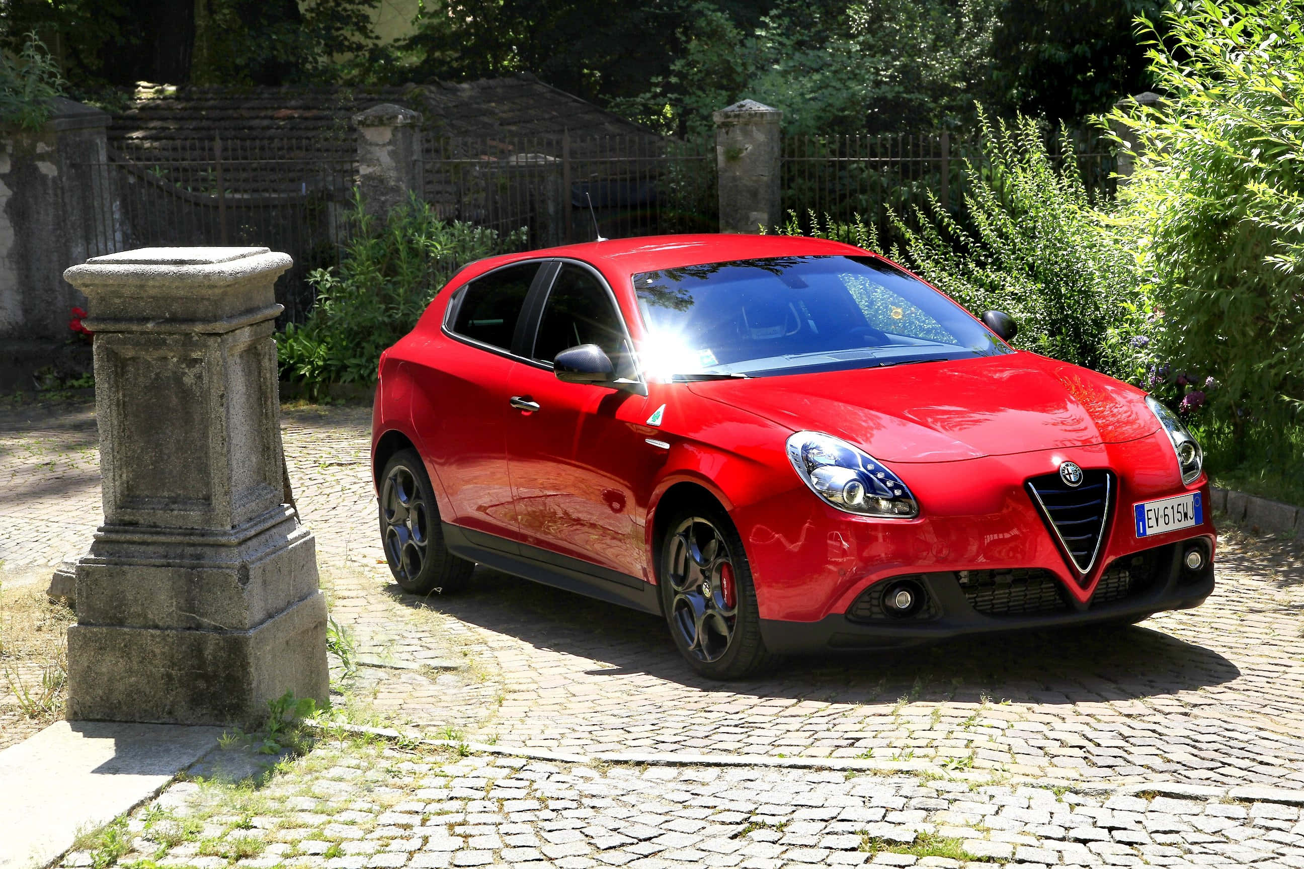 Alfa Romeo Giulietta 桌布 壁紙 — 一輛鮮紅色的愛快羅密歐Giulietta停放在陽光明媚的鵝卵石車道上，旁邊是一根風化的石柱。