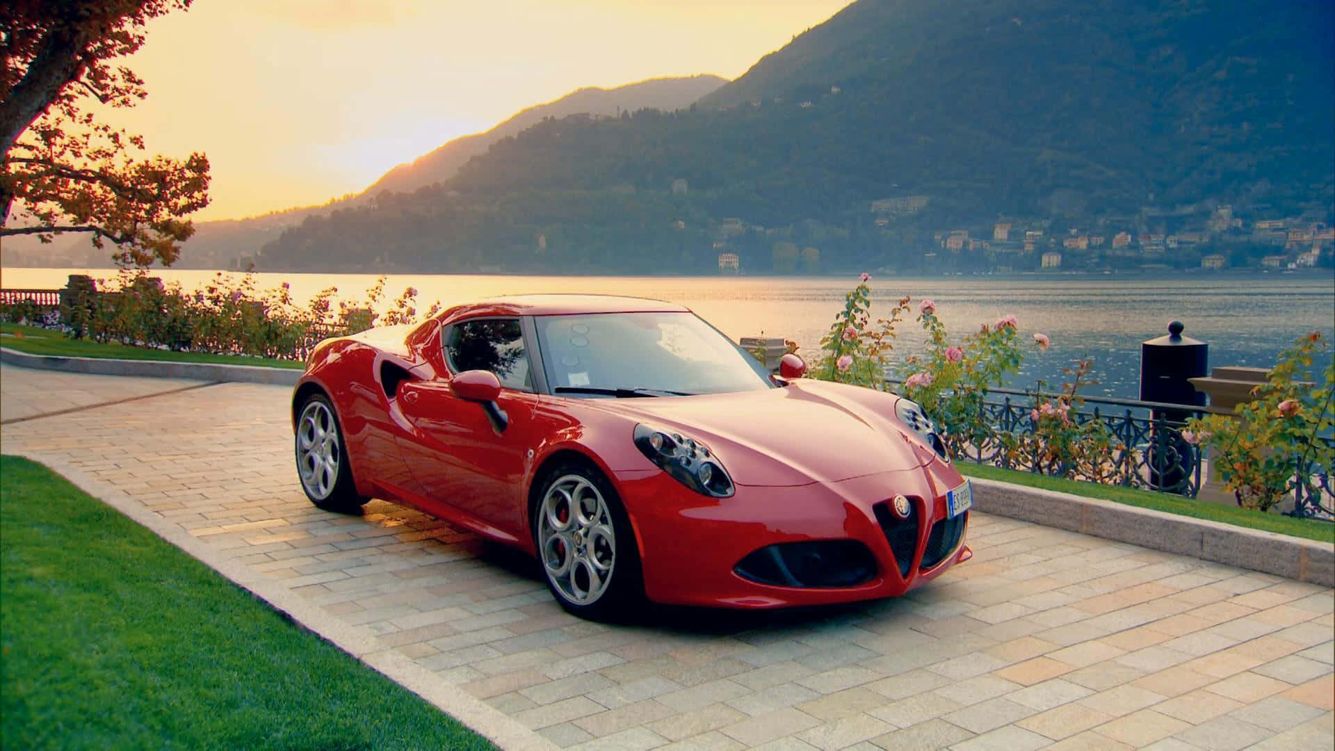 Alfa Romeo 4c 桌布 壁紙 — 一輛鮮紅色的愛快羅密歐跑車停泊在寧靜湖泊旁的石徑上，背景是金色夕陽和群山。