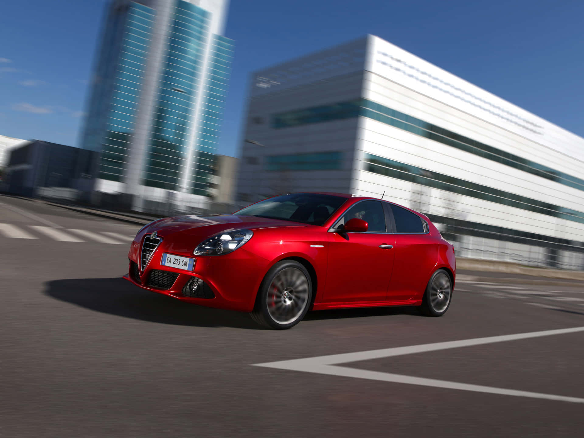 Alfa Romeo Giulietta 桌布 壁紙 — 一輛鮮紅色的愛快羅密歐跑車在晴朗藍天下，駛過現代都市街道，兩旁是模糊的玻璃摩天大樓。