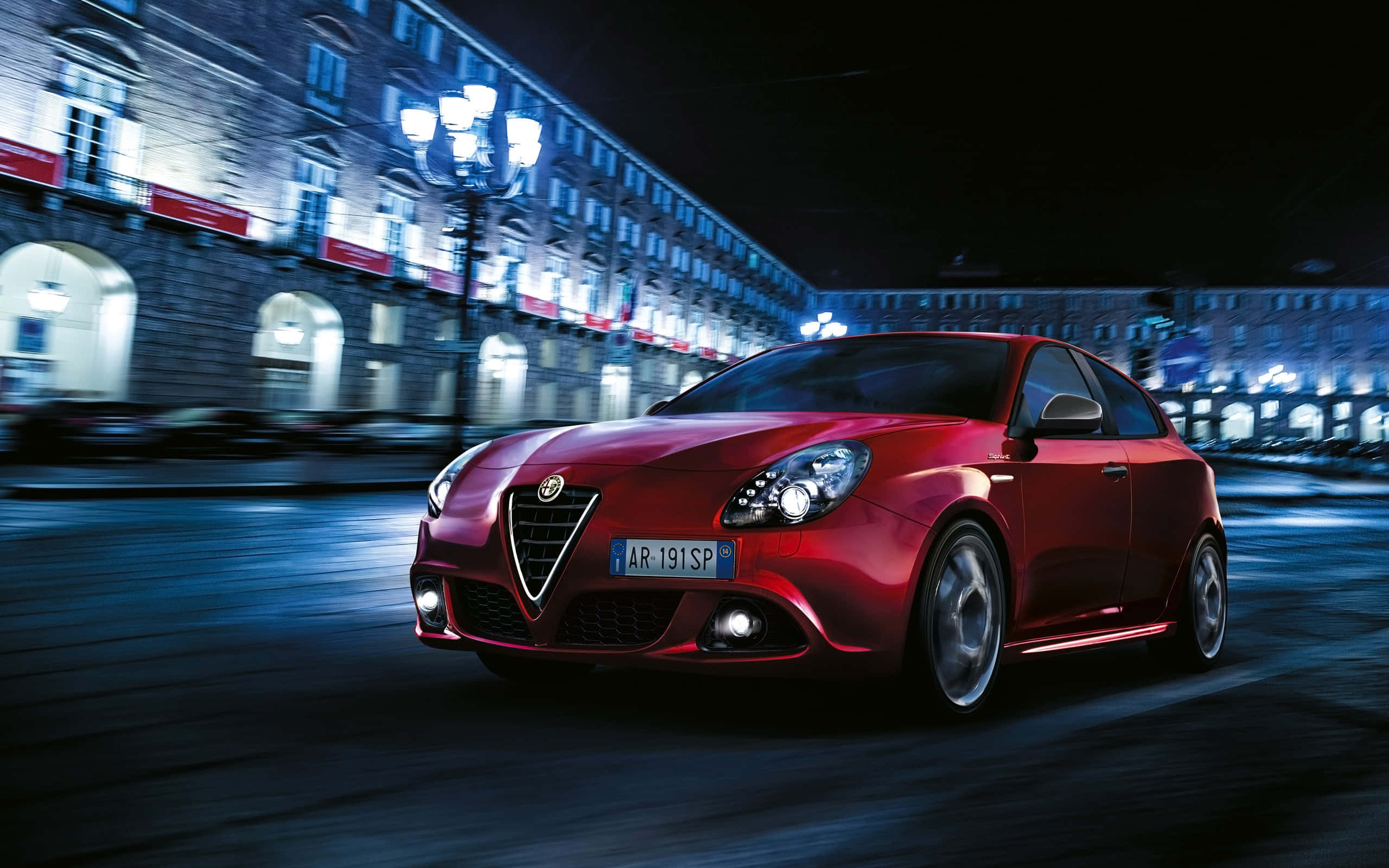 Alfa Romeo Giulietta 桌布 壁紙 — 一輛鮮紅色的愛快羅密歐跑車在模糊的夜間城市街道上飛馳，街道被閃爍的路燈和建築物外牆照亮。