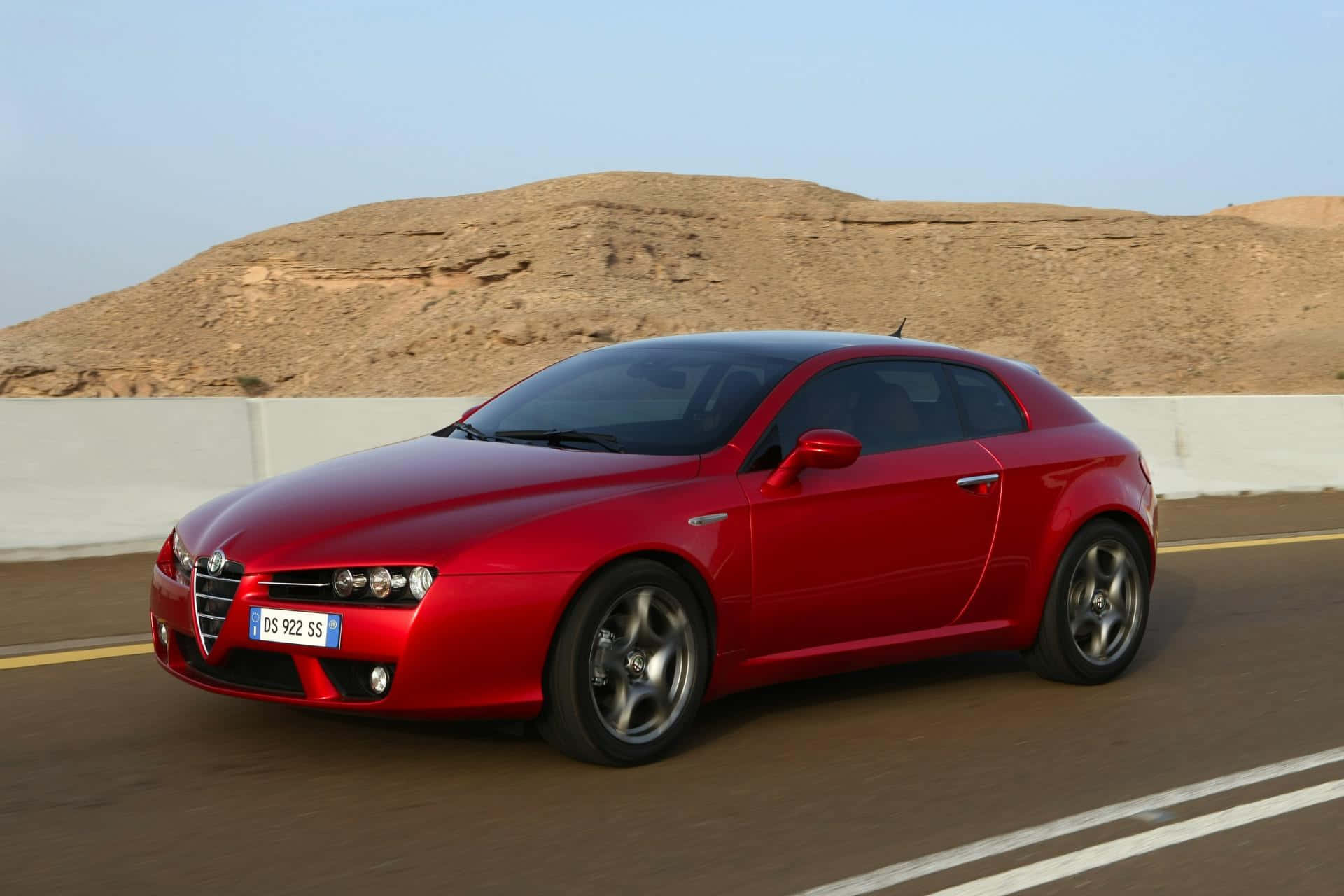 Alfa Romeo Brera 桌布 壁紙 — 一輛鮮紅色的愛快羅密歐跑車在沙漠公路上行駛，背景是沙丘。