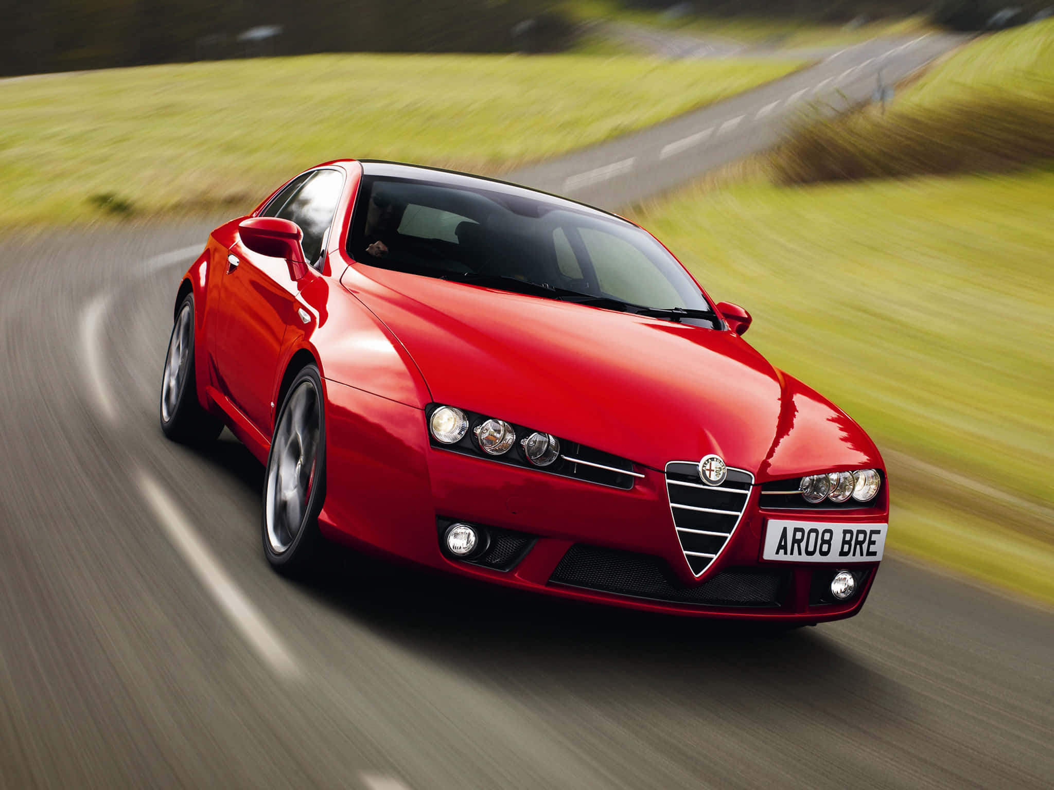 Alfa Romeo Brera 桌布 壁紙 — 一輛鮮紅色的愛快羅密歐跑車在蜿蜒的鄉村道路上疾馳，背景是模糊的綠色田野和瀝青路面。