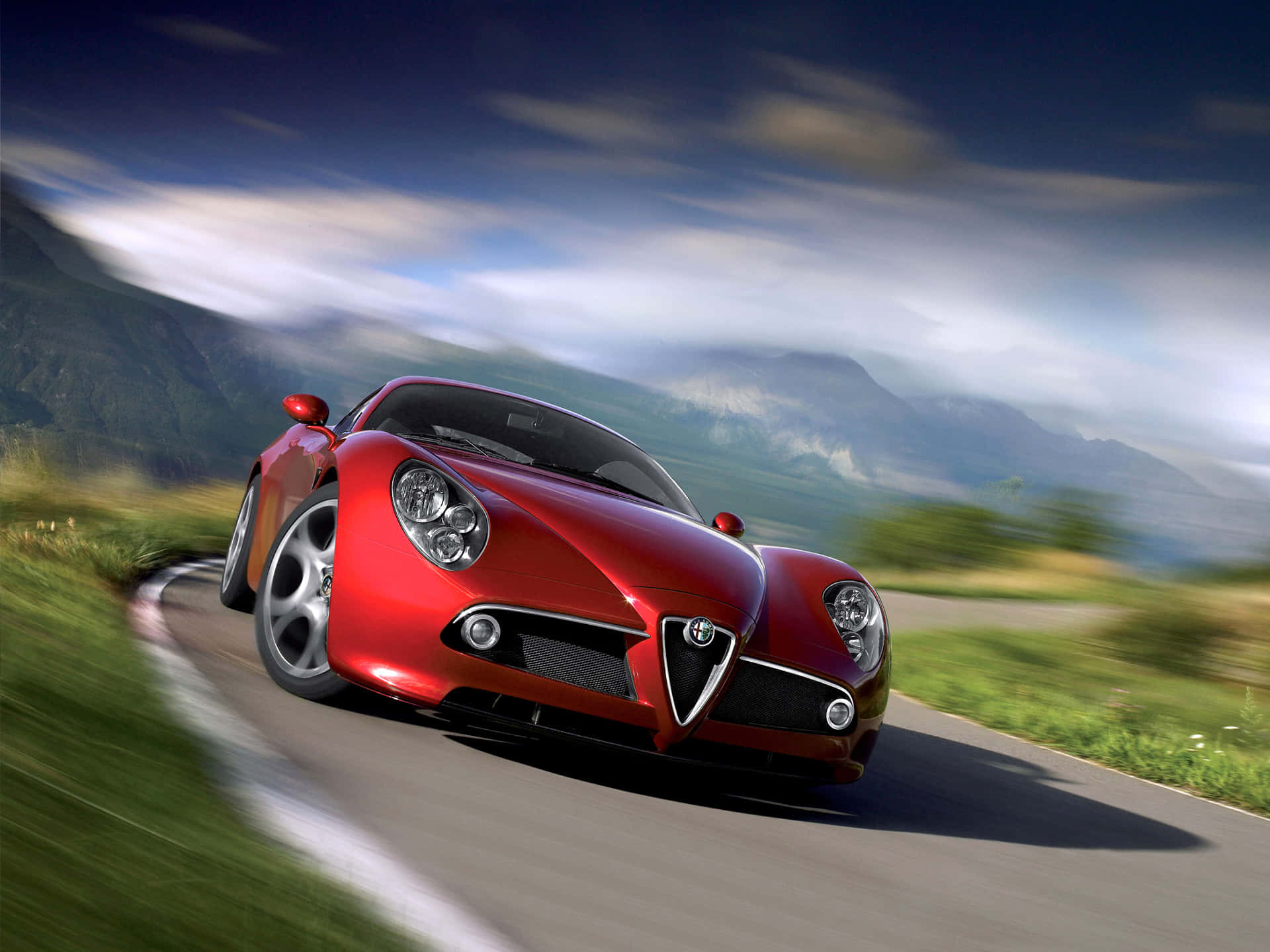 Alfa Romeo 8c Competizione 桌布 壁紙 — 一輛鮮紅色的愛快羅密歐跑車在蜿蜒的山路上疾馳，背景是模糊的綠意和遠處的山峰，天空壯麗。