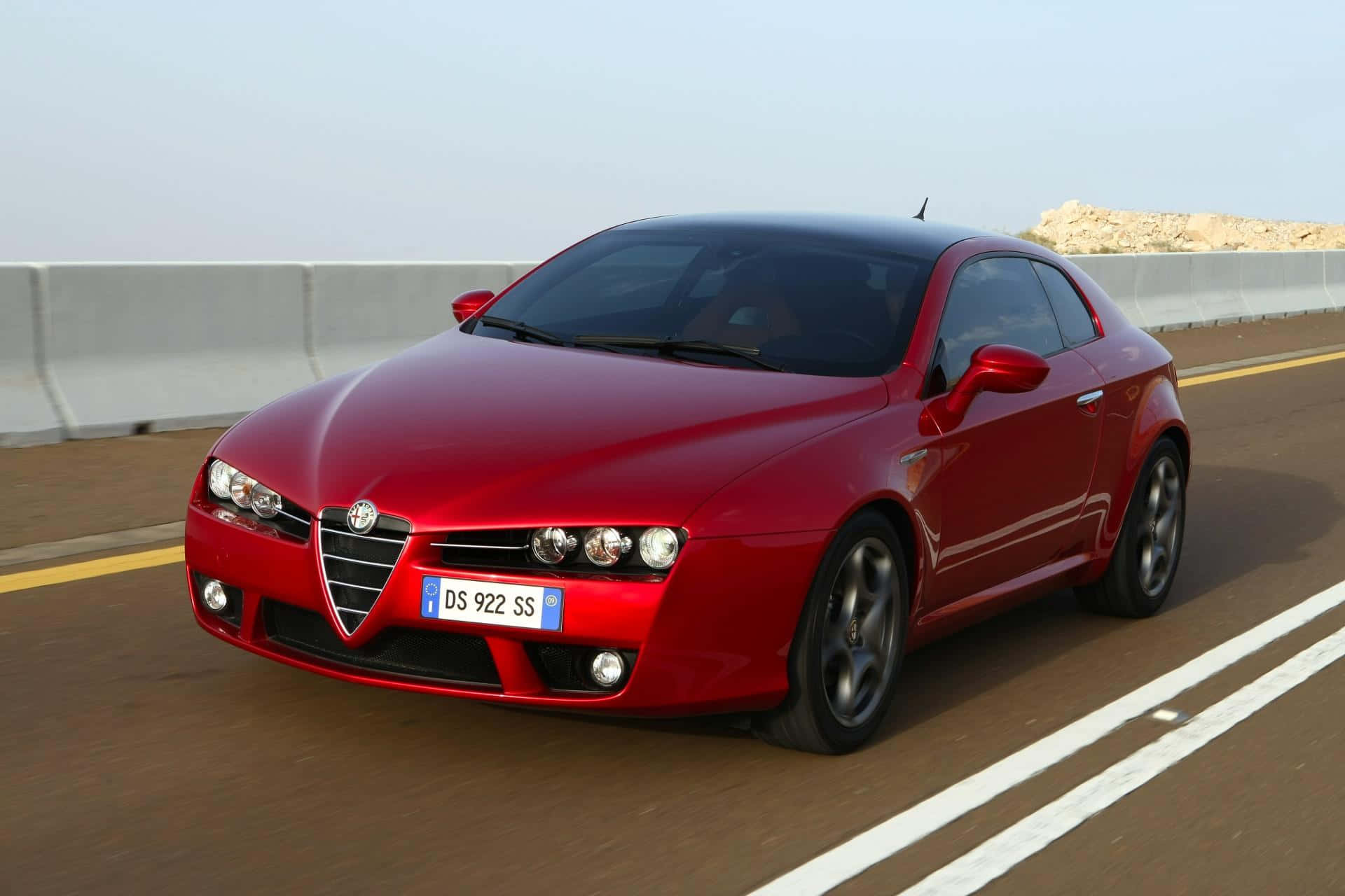 Alfa Romeo Brera 桌布 壁紙 — 一輛鮮紅色的愛快羅密歐跑車在陽光明媚的高速公路上行駛，背景有混凝土護欄和岩石地形。