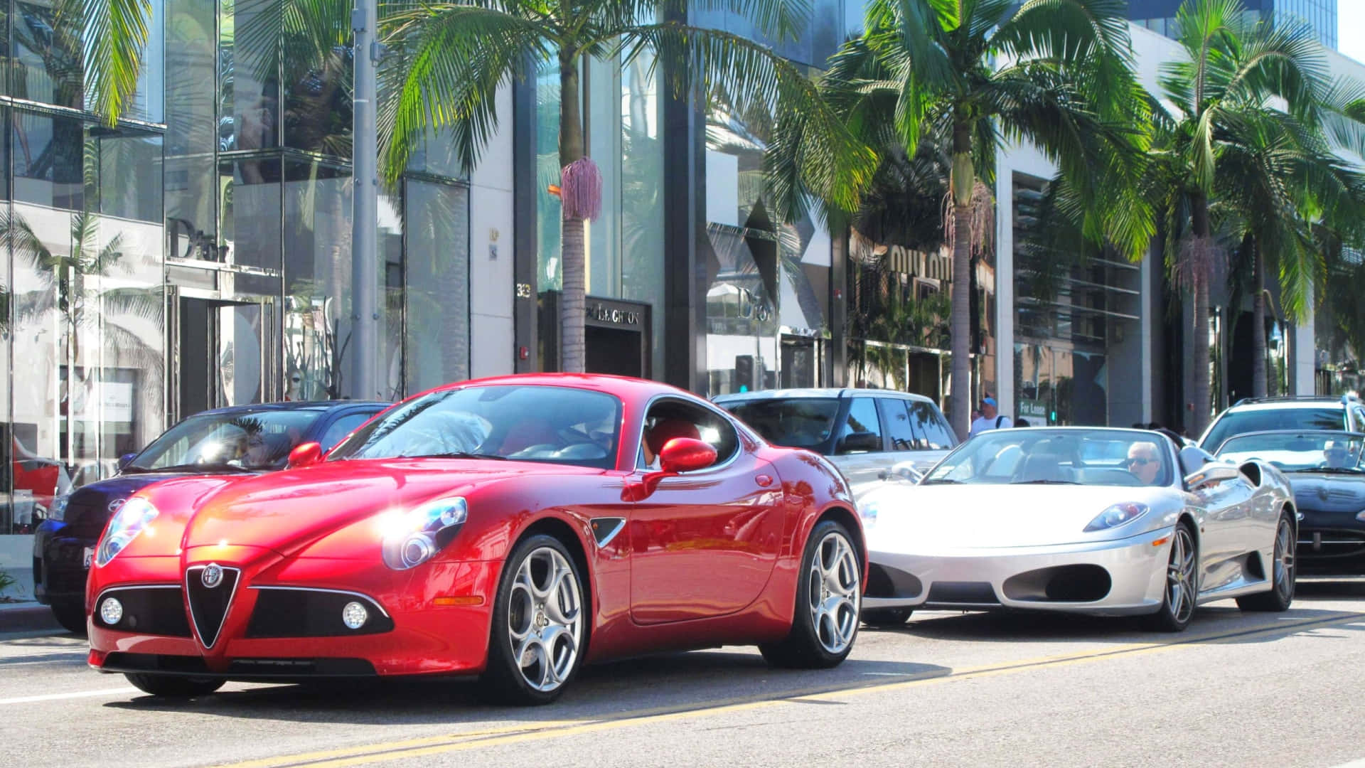 Alfa Romeo 8c Competizione 桌布 壁紙 — 一輛鮮紅色的愛快羅密歐跑車帶領一排豪華車輛，行駛在棕櫚樹成蔭的城市街道上，陽光明媚。