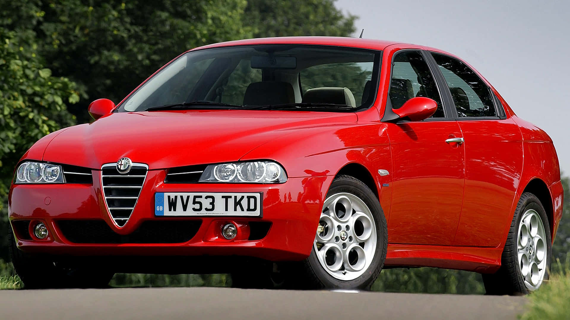 Alfa Romeo 156 桌布 壁紙 — 一輛鮮紅色的愛快羅密歐轎車在日光下閃閃發光，停泊在一條背景有茂密綠樹的道路上。