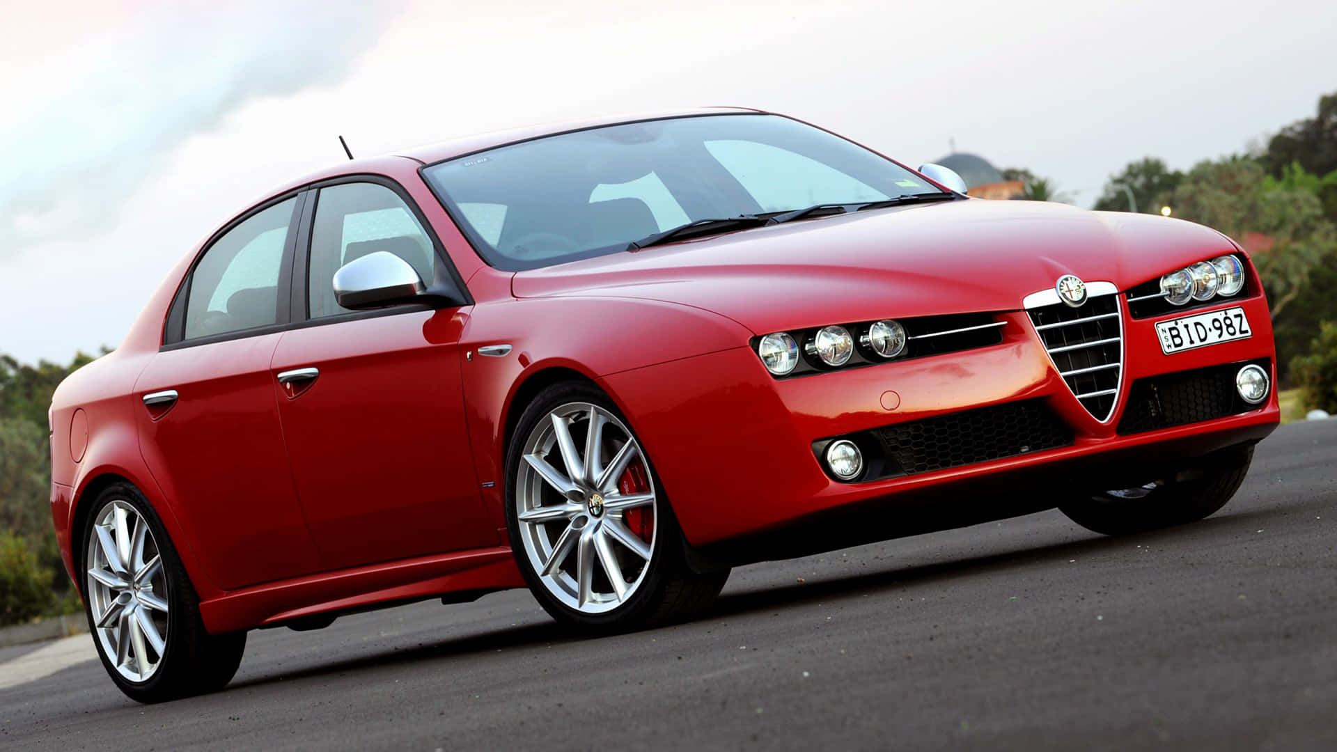 Alfa Romeo 159 桌布 壁紙 — 一輛鮮紅色的愛快羅密歐轎車在柔和日光下閃閃發光，停在鋪砌的道路上，背景有綠化。