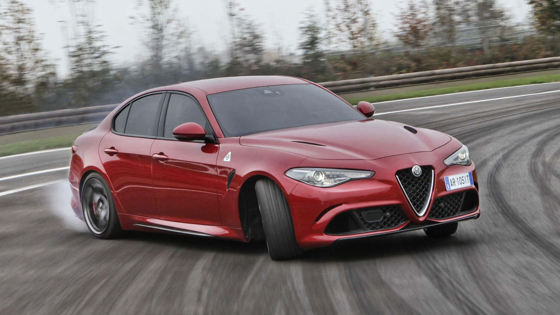 Alfa Romeo Giulia 桌布 壁紙 — 一輛鮮紅色的愛快羅密歐轎車在潮濕的賽道上甩尾，後輪冒出煙霧，周圍樹木環繞。