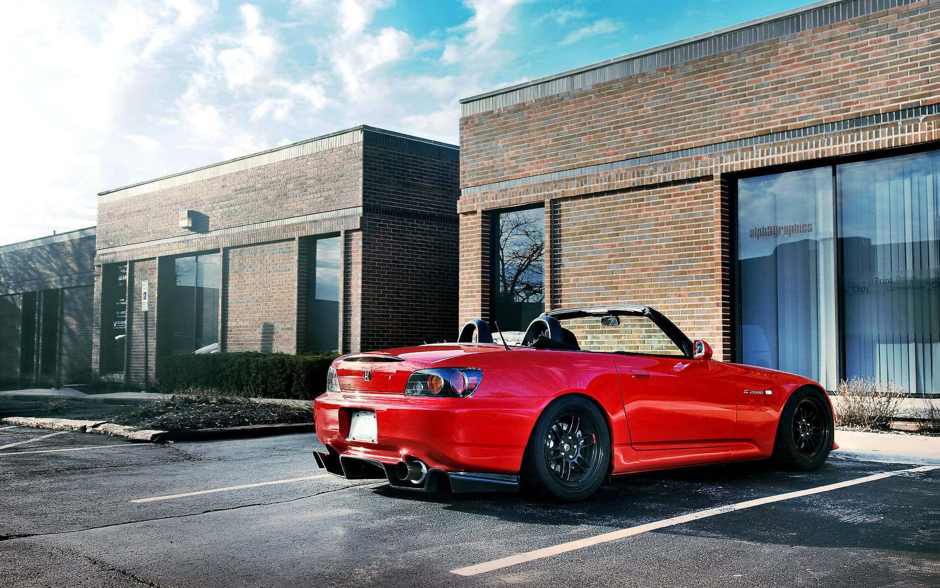 Honda S2000 桌布 壁紙 — 一輛鮮紅色的敞篷跑車停放在磚造建築旁的停車場，上方是晴朗的藍天，點綴著幾朵白雲。