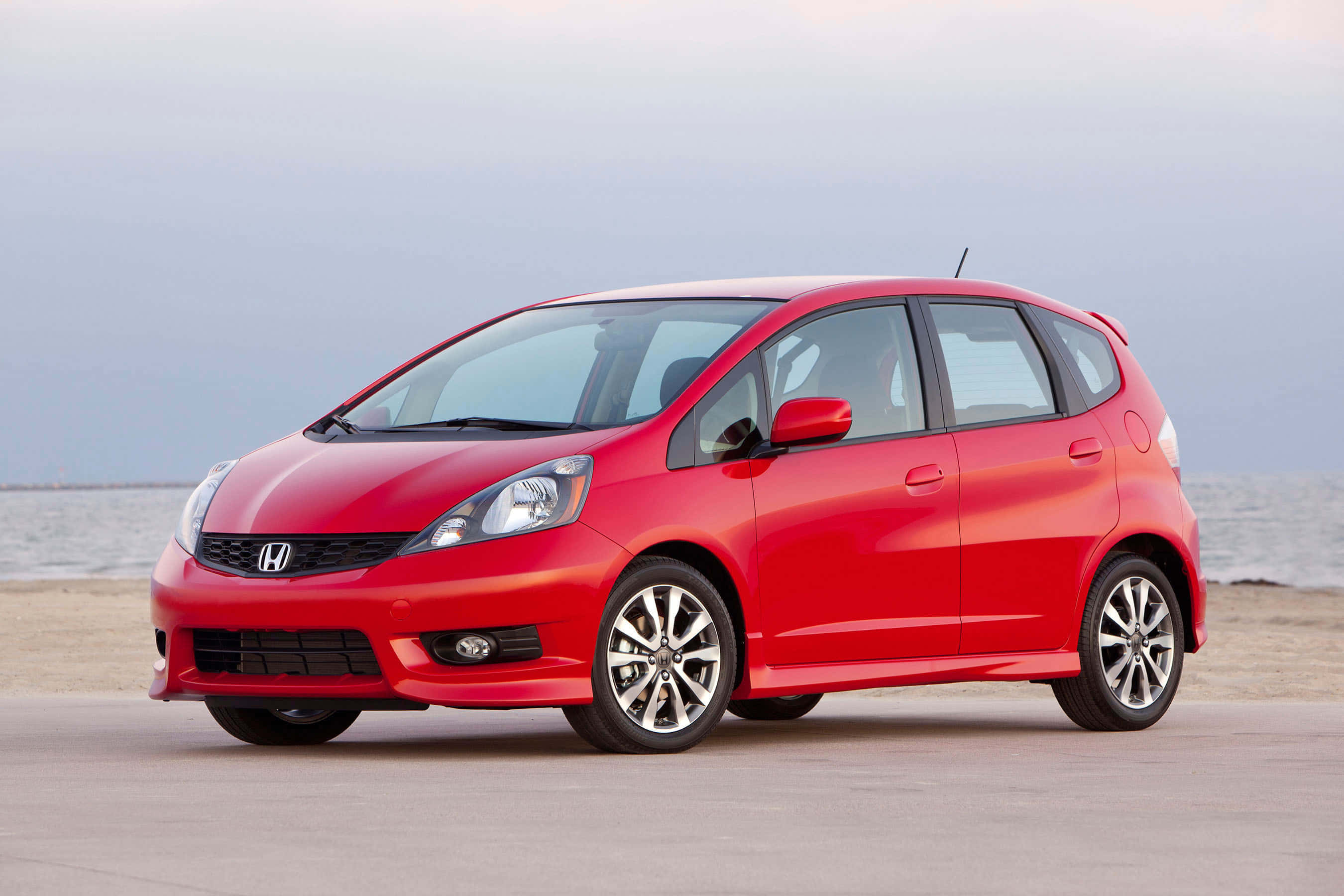 Honda Fit 桌布 壁紙 — 一輛鮮紅色的本田Fit停泊在沙灘上，背景是海洋和多雲的天空。