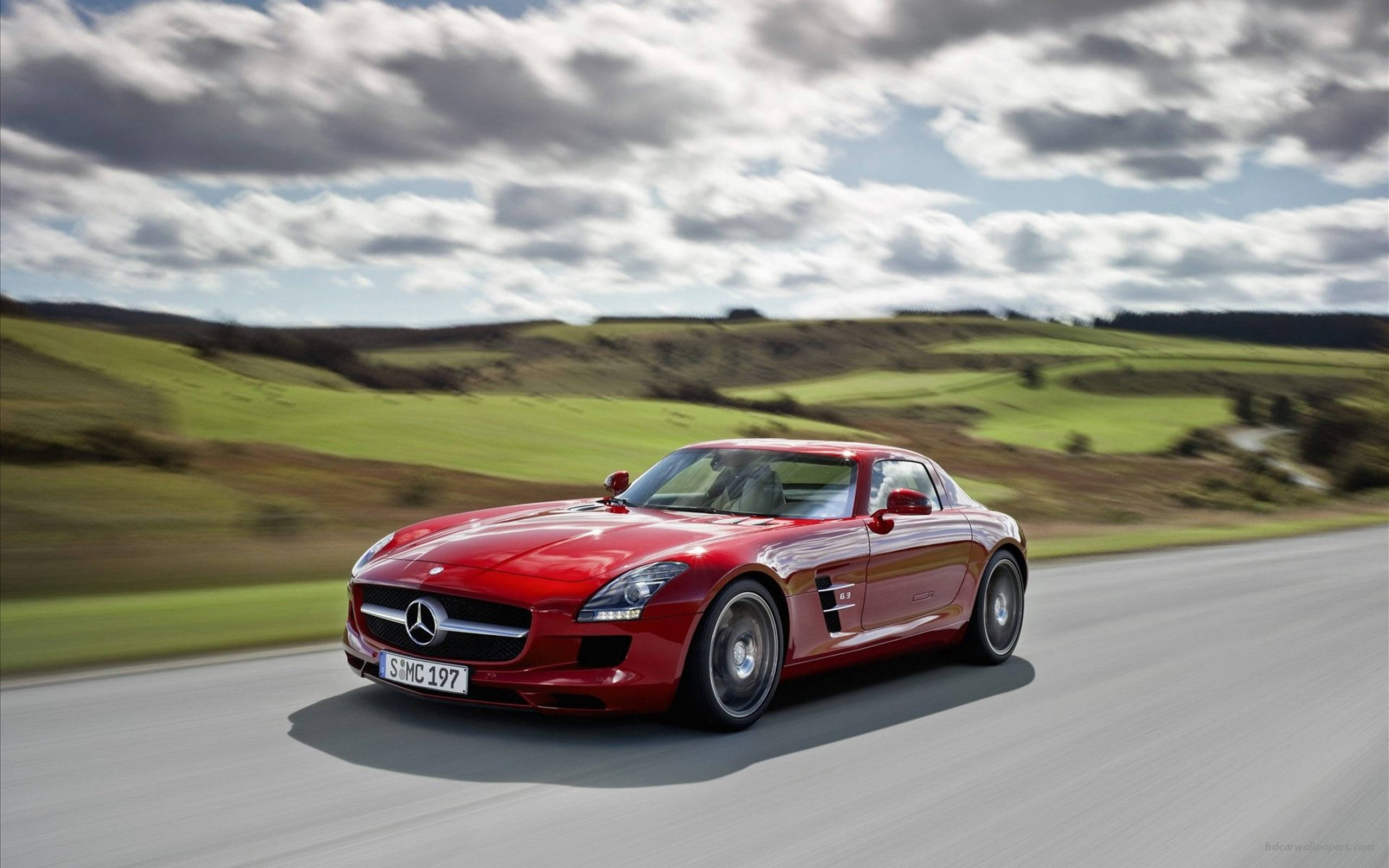 高清 桌布 壁紙 — 一輛鮮紅色的梅賽德斯 SLS AMG 跑車在陰天之下，沿著蜿蜒的道路穿過鬱鬱蔥蔥的綠色山丘飛馳。